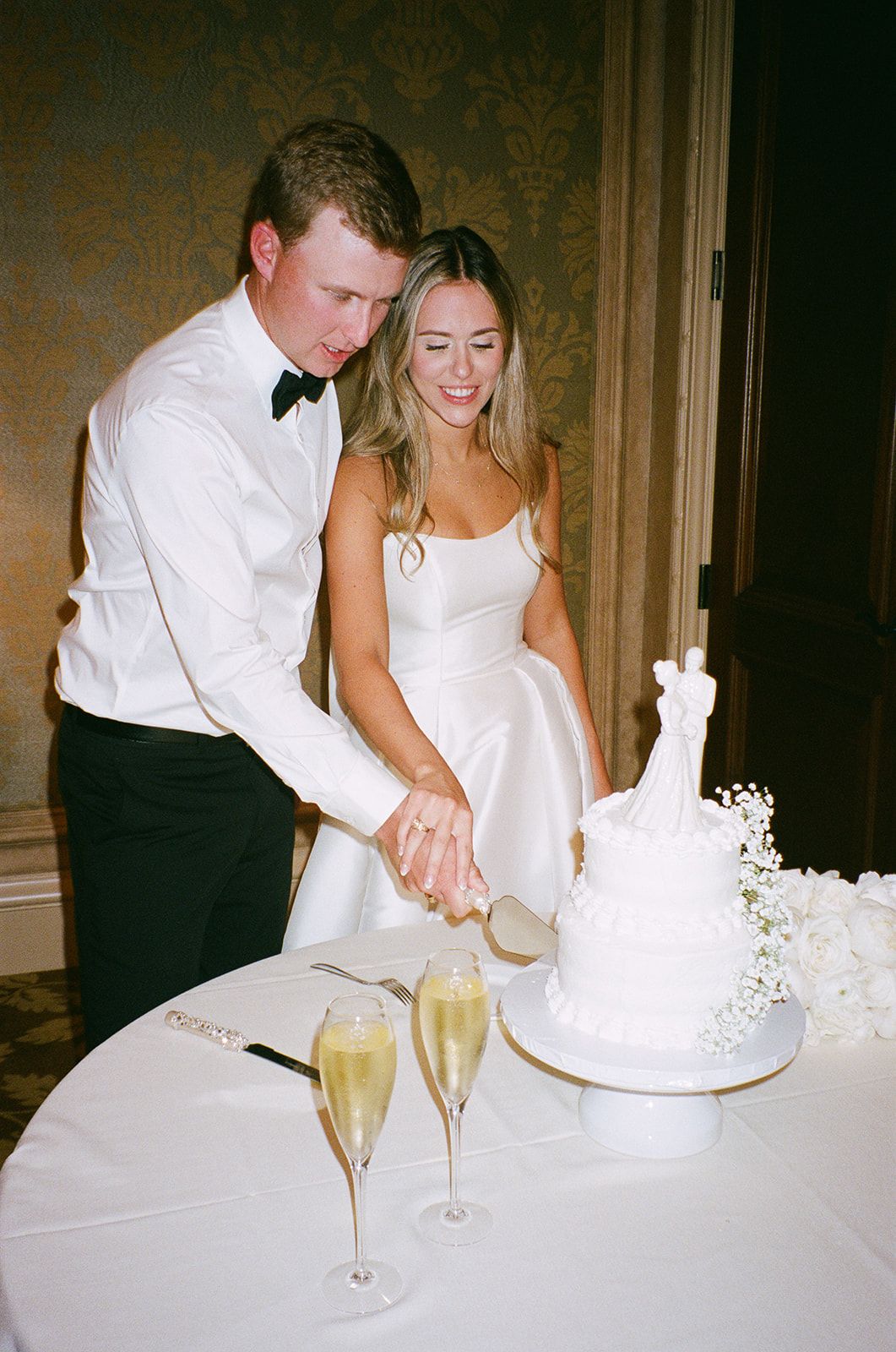 Couple in white attire cutting their wedding cake together at the reception.