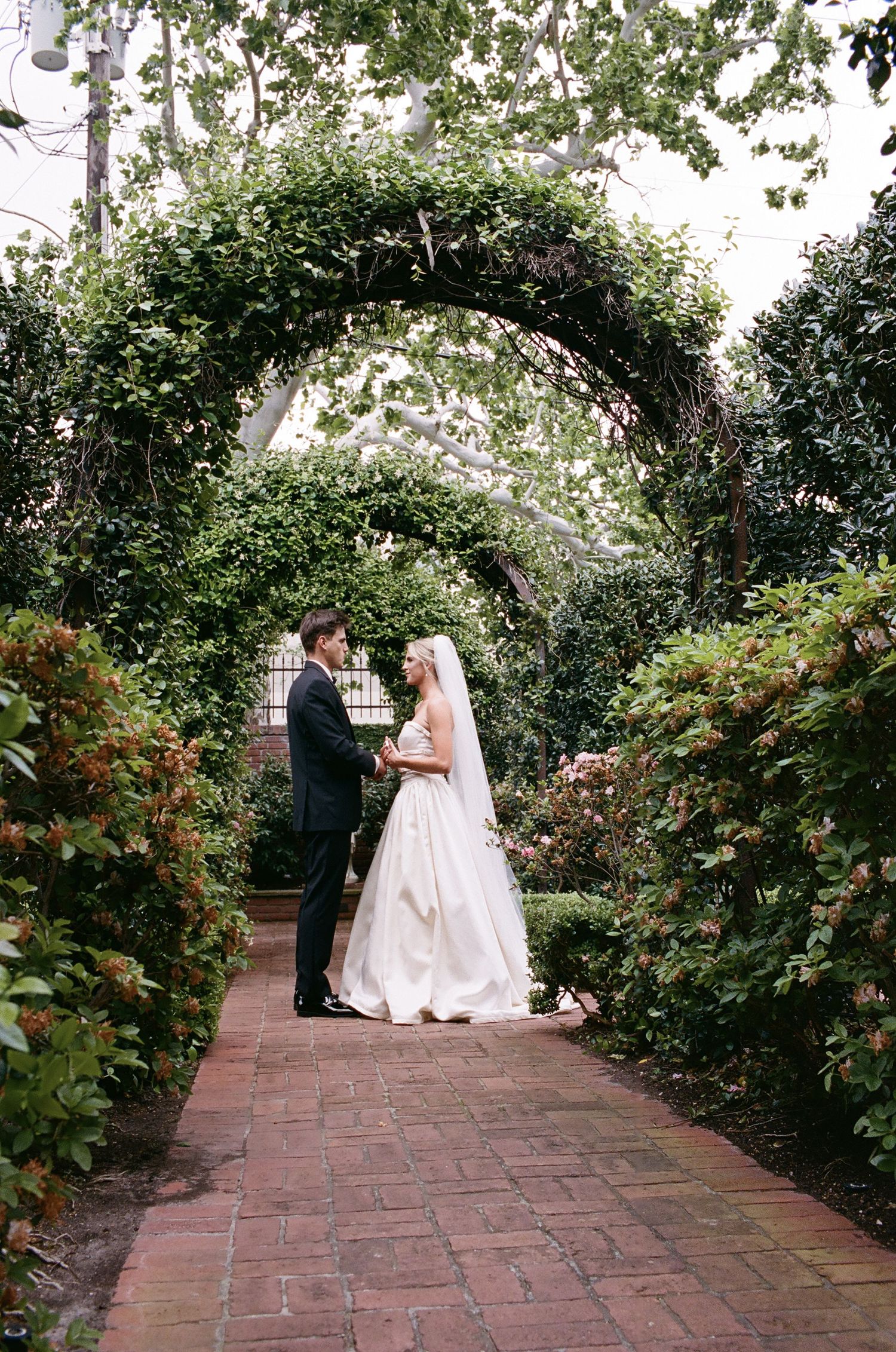 Bride and groom stand under lush green garden archway on brick path surrounded by flowers.