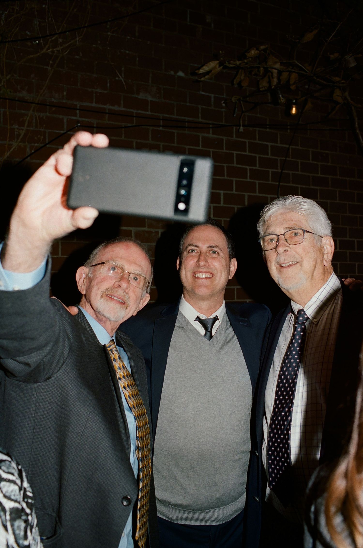 Three people posing together while one takes a selfie photo with a mobile phone at an indoor gathering.