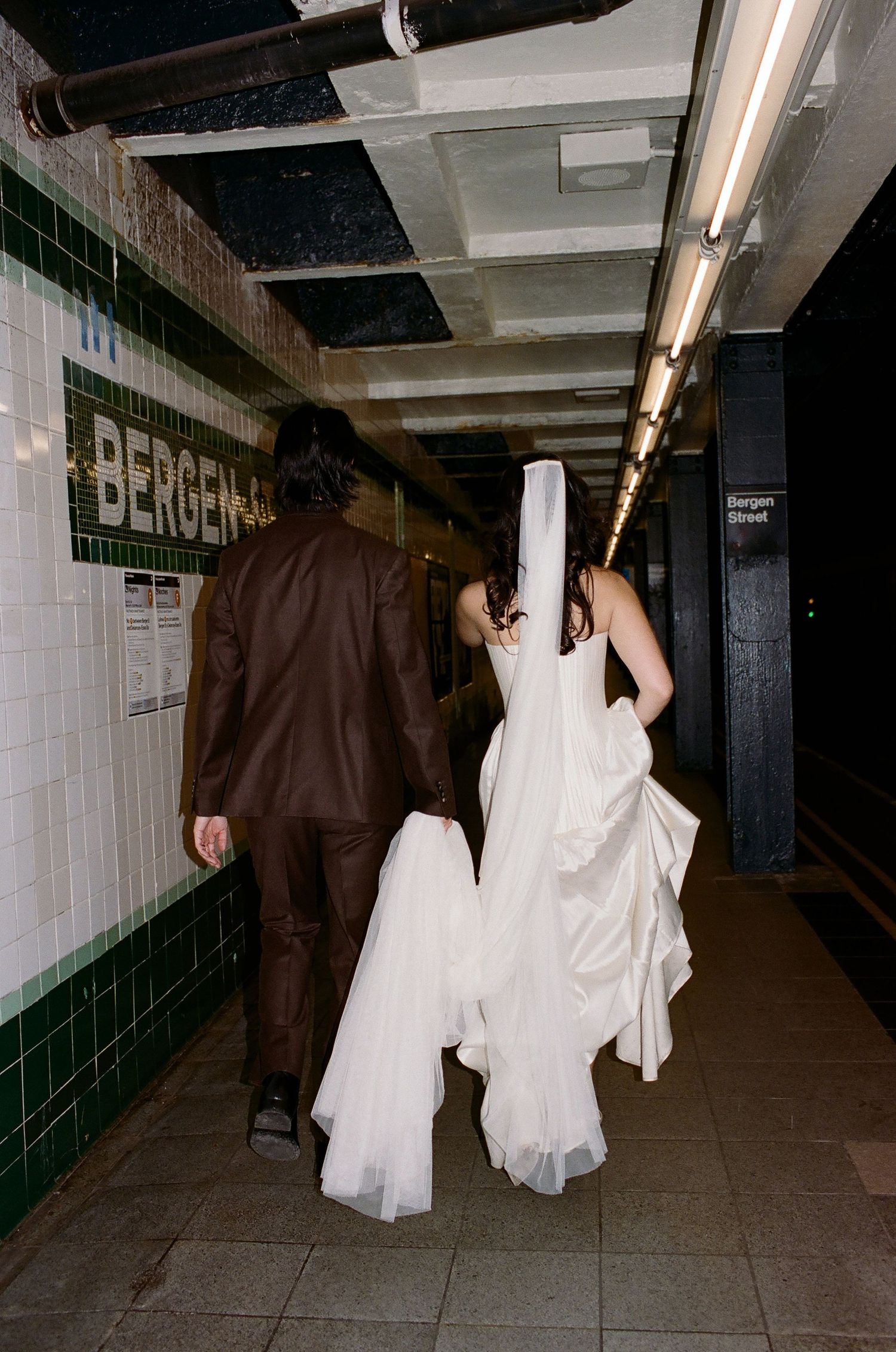 Two people in contrasting white and brown outfits walk through a dimly lit subway tunnel with tiled walls.