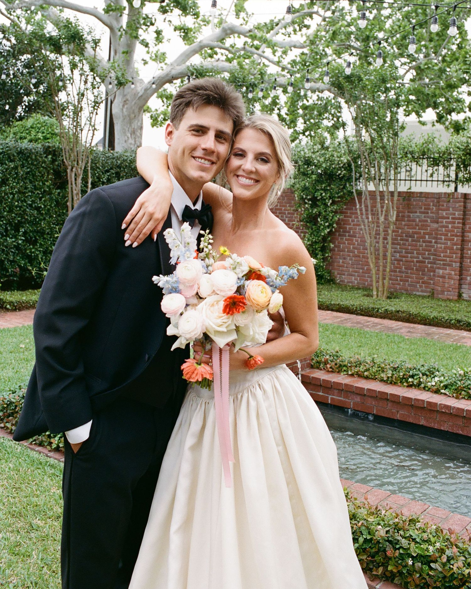 A wedding couple poses in a garden setting with a brick wall and water feature in the background.