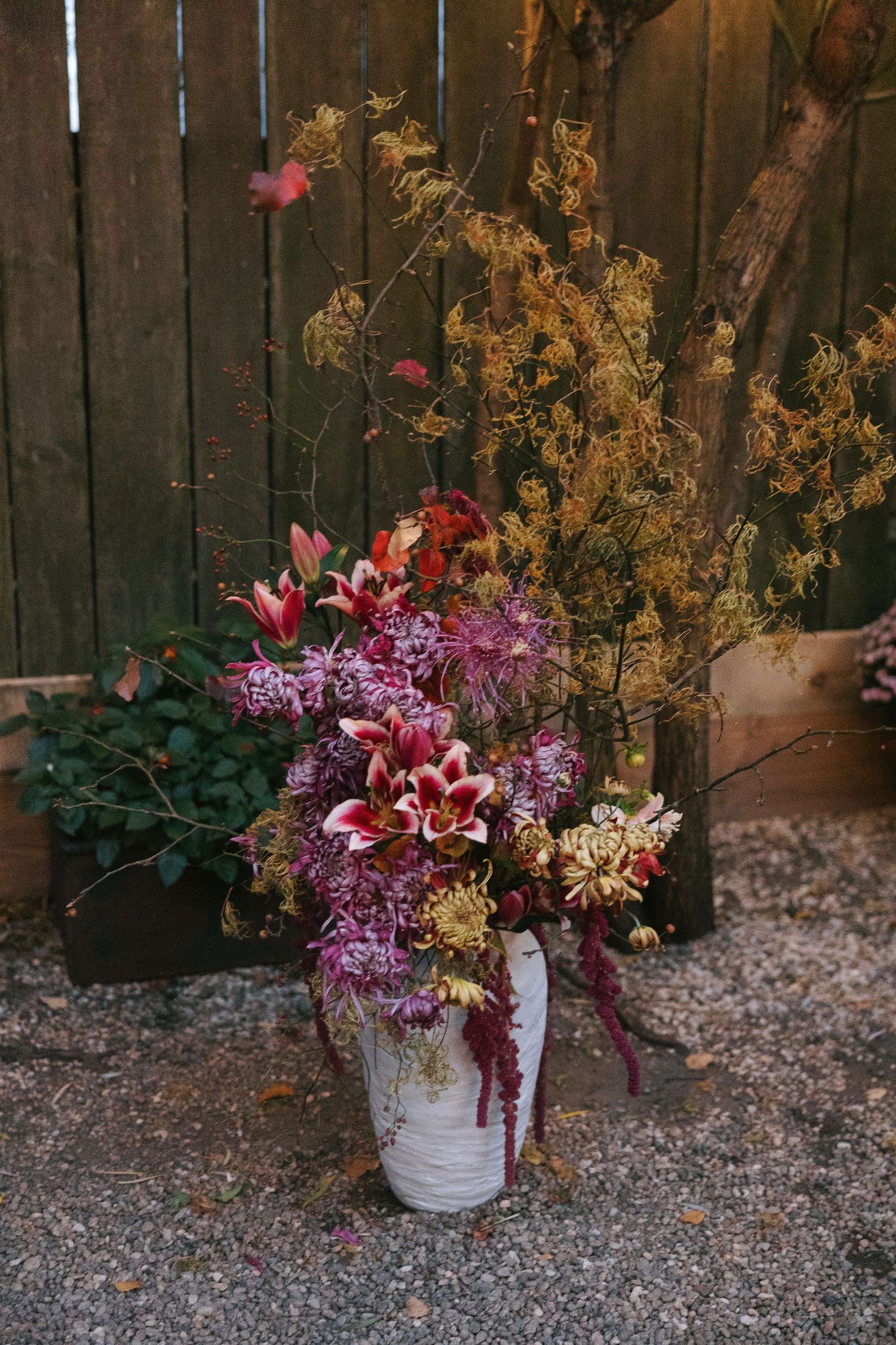 A rustic floral arrangement with pink lilies and dried flowers in a white vase against a wooden fence.