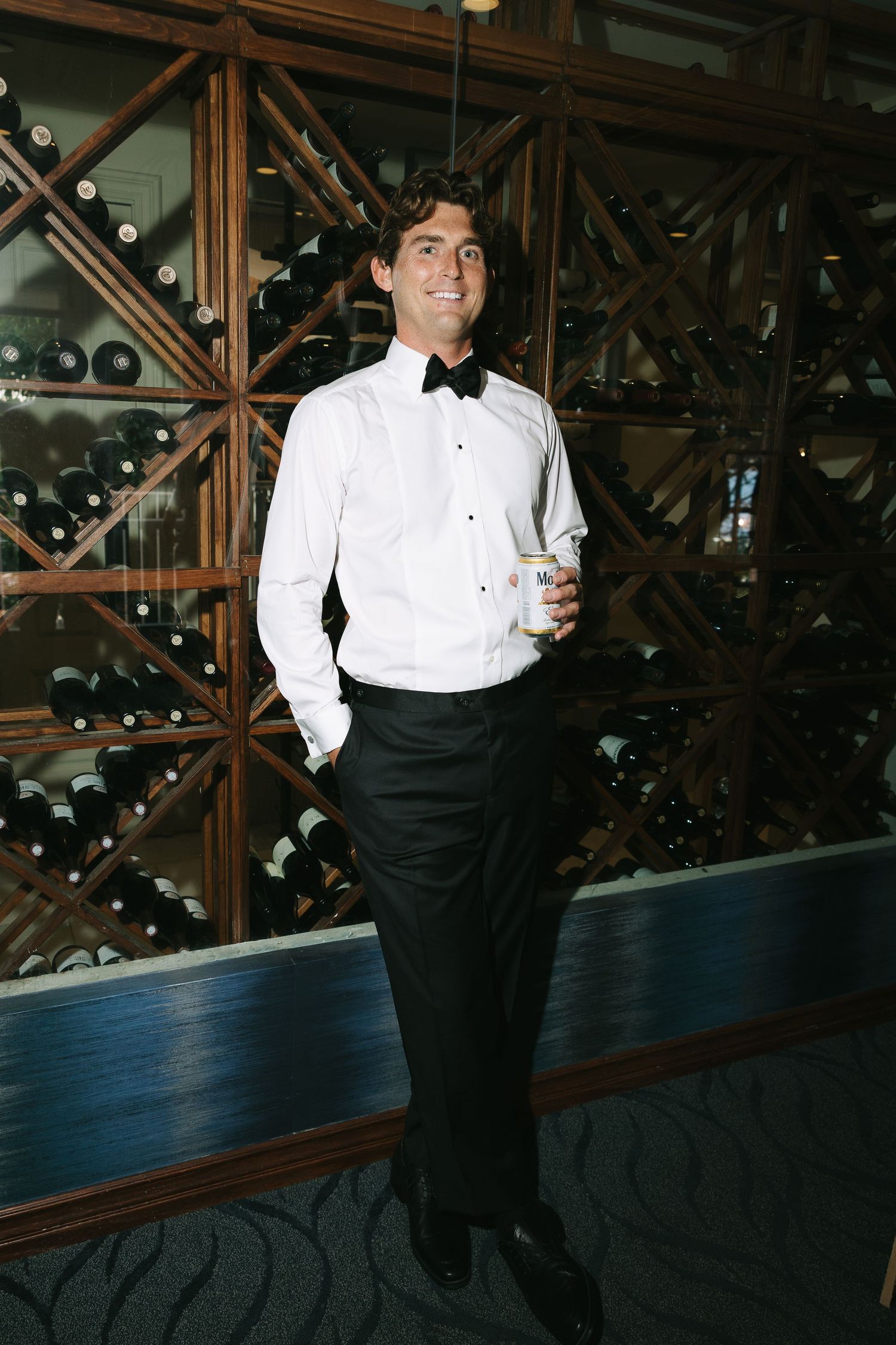 A person in formal wear stands against wooden wine racks in an elegant cellar setting.
