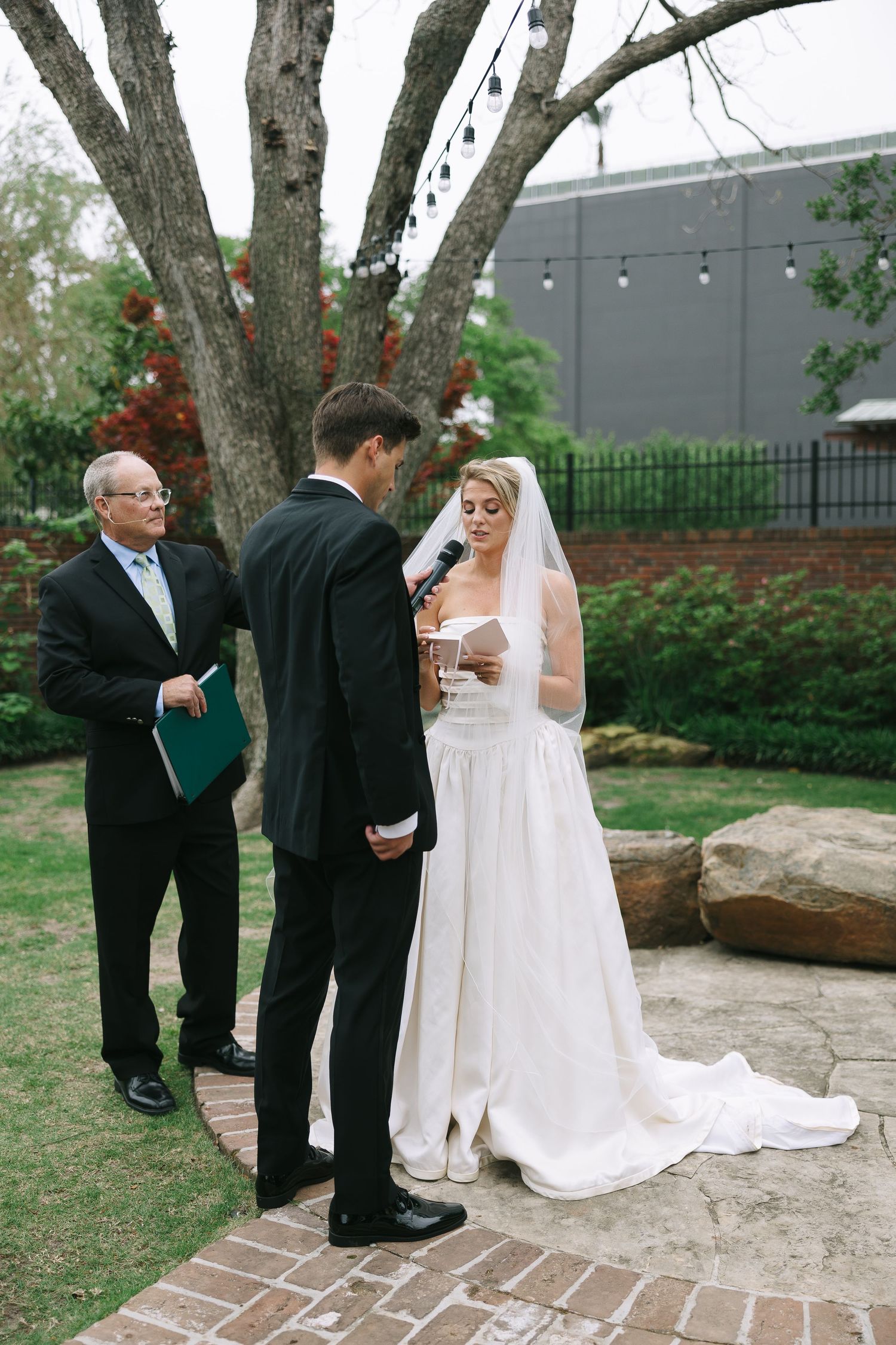 Outdoor wedding ceremony on brick pathway with trees and red flowers in background.