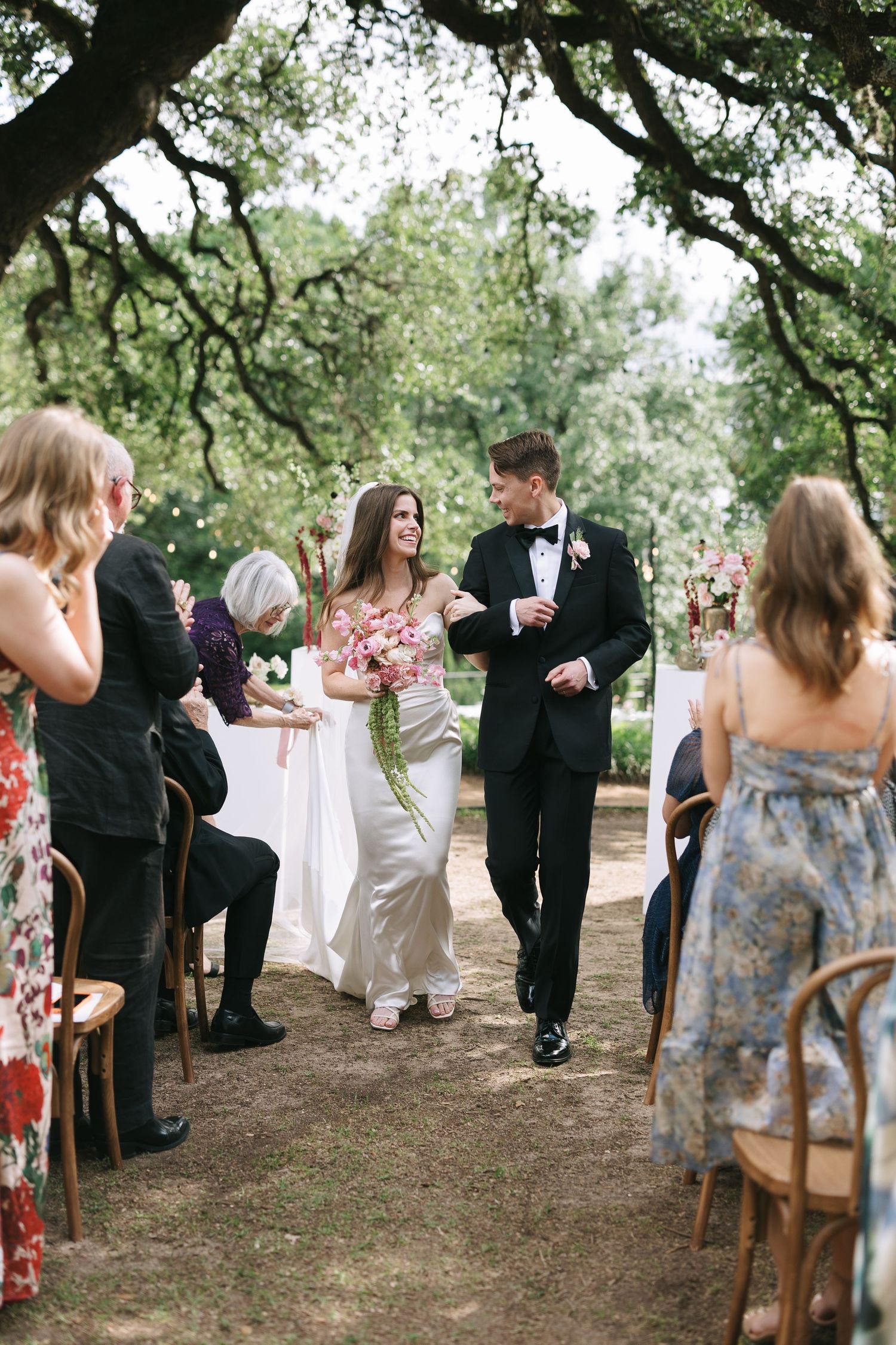 Wedding guests seated on wooden chairs during an outdoor ceremony beneath sprawling oak trees.
