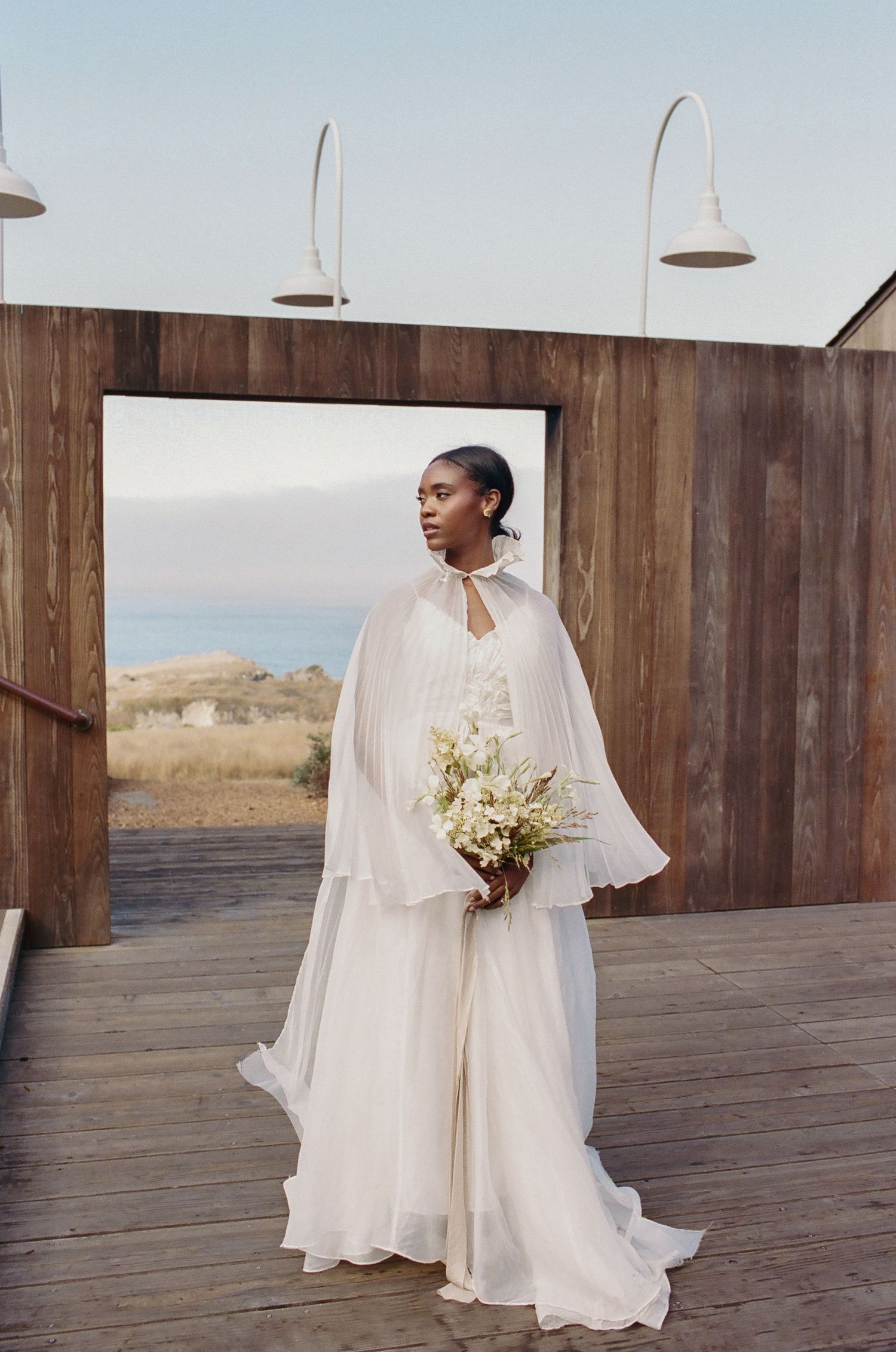 Elegant bride in white flowing gown and cape holds bouquet on wooden deck with ocean view backdrop.