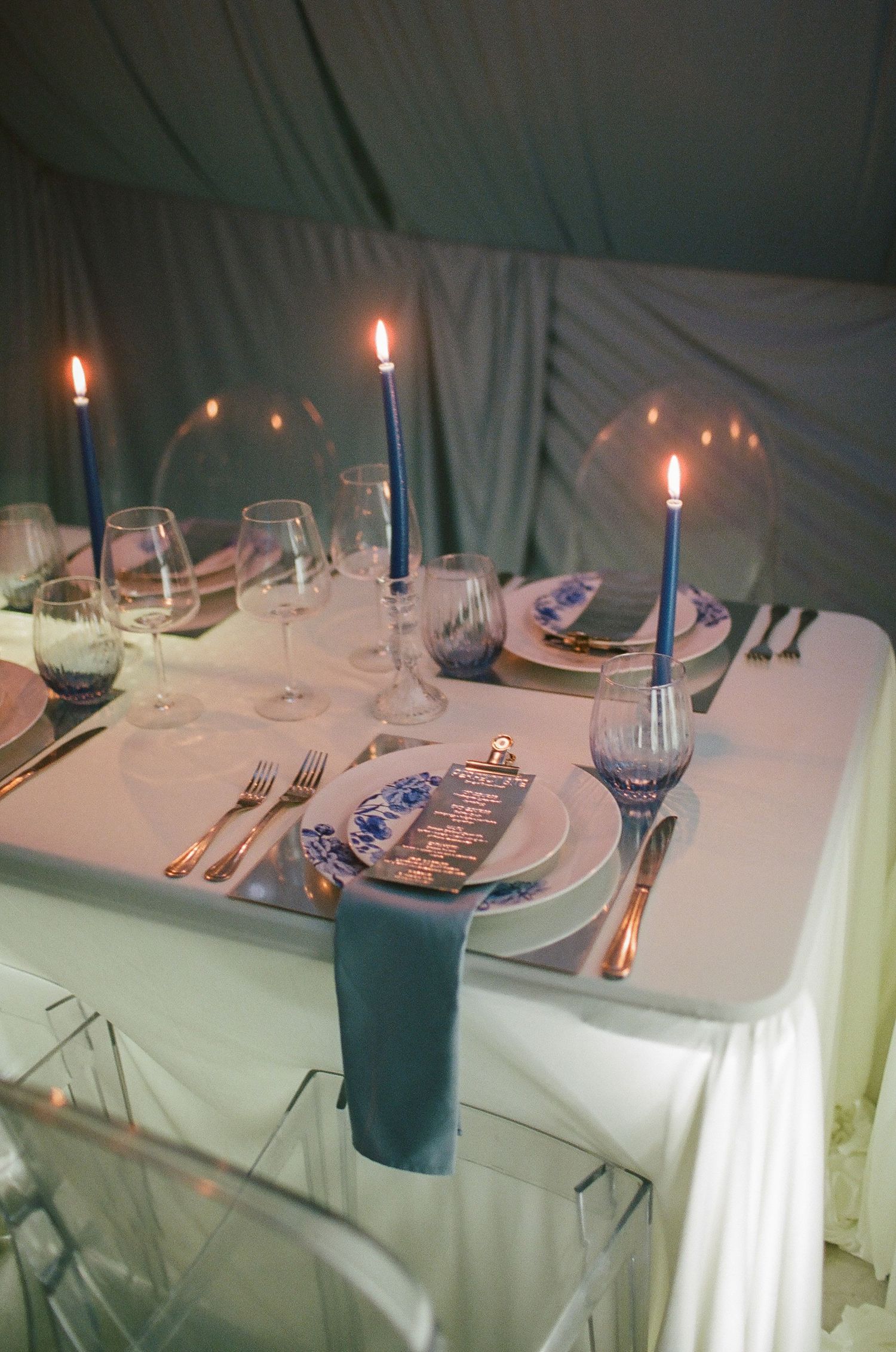 Elegant table setting with blue taper candles, white linens, blue napkins, and blue and white plates with gold cutlery.