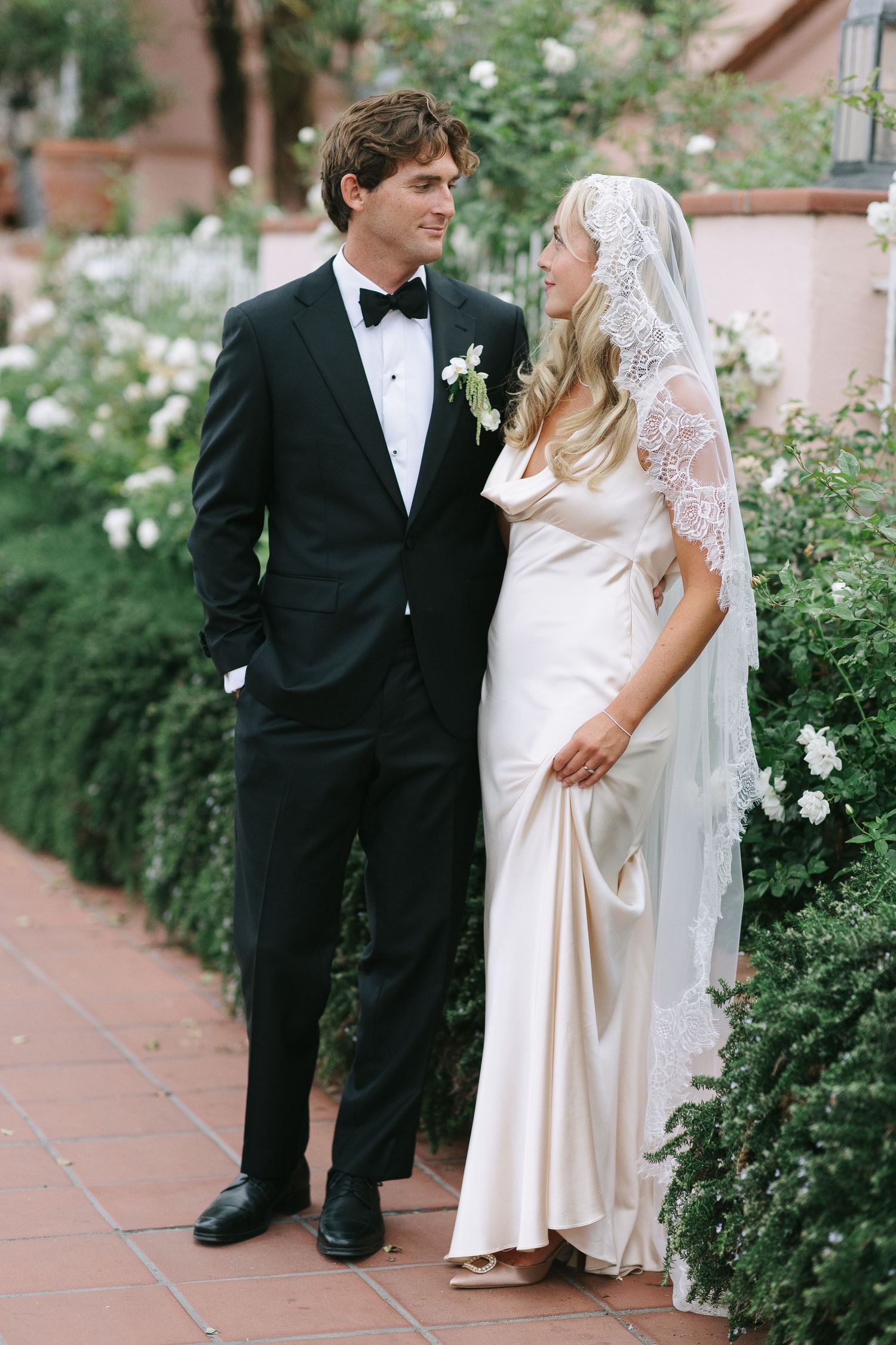 Newlyweds share intimate moments in a beautiful garden setting with white roses and brick pathways.