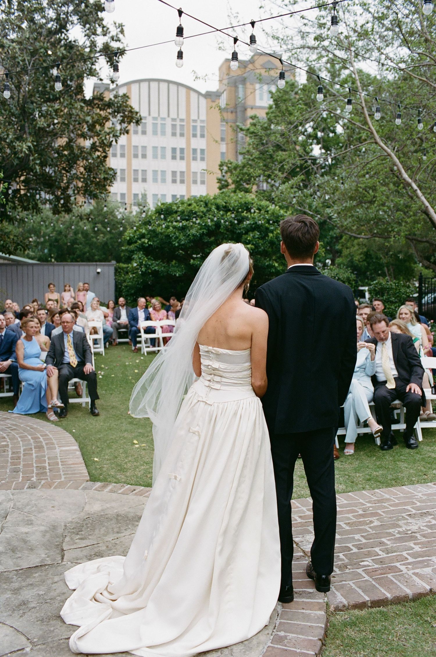 Outdoor garden wedding ceremony with string lights and guests seated on white chairs.