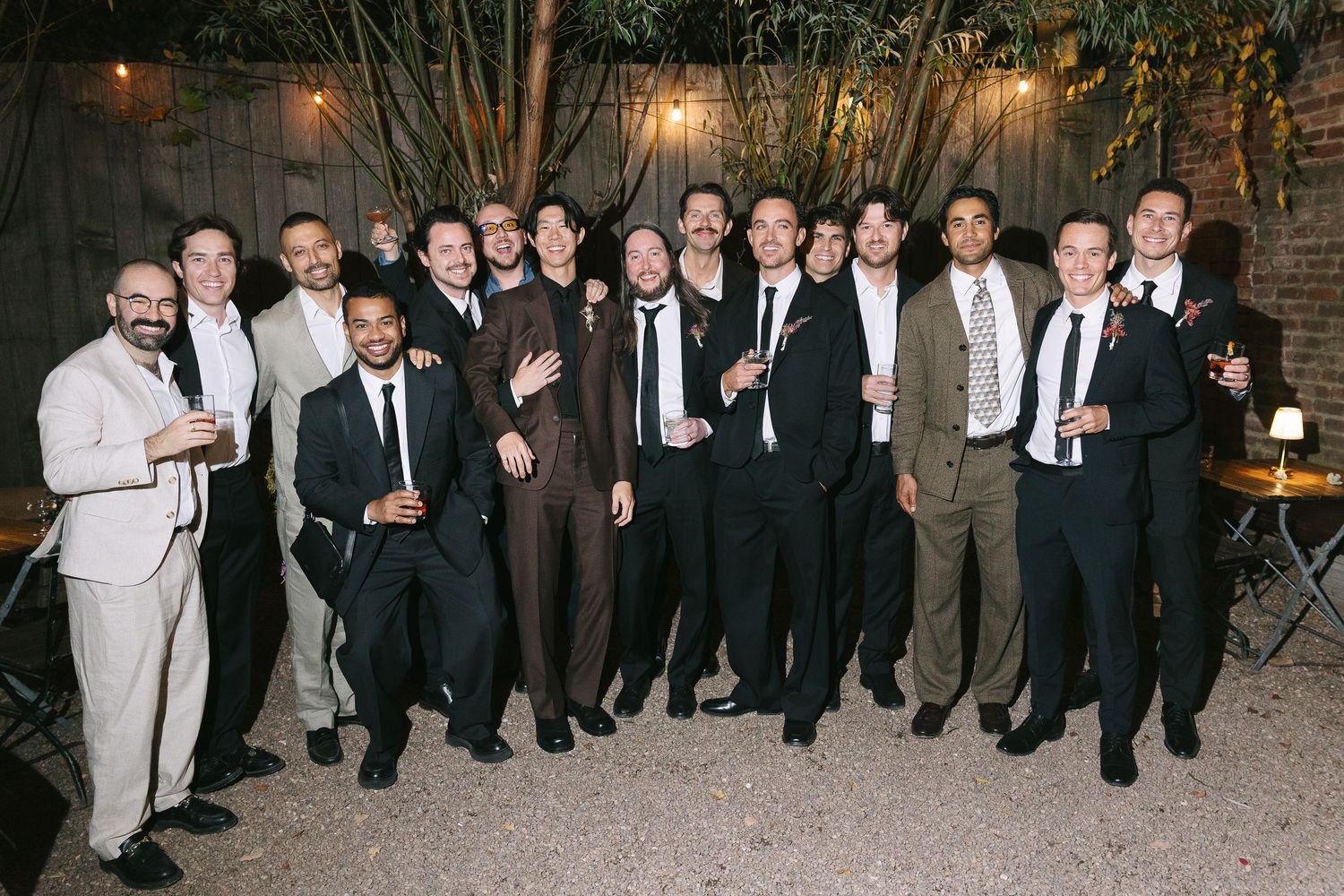 Groomsmen and wedding guests in formal attire gather for a group photo at an outdoor reception.