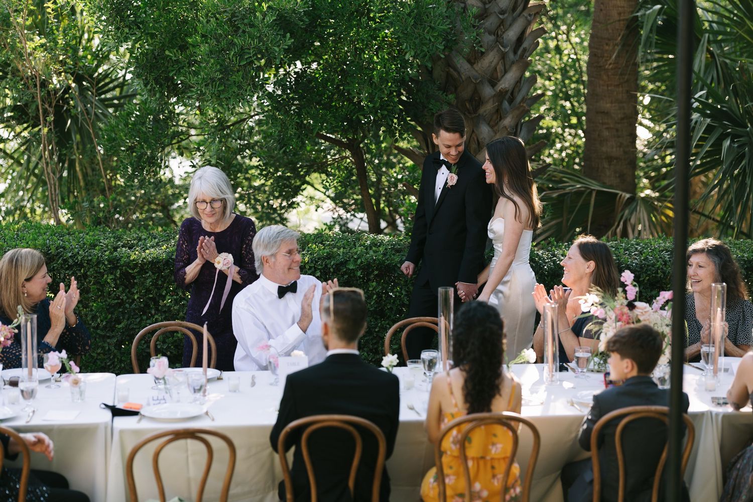 Wedding guests seated at long tables with wooden chairs in an outdoor garden setting.