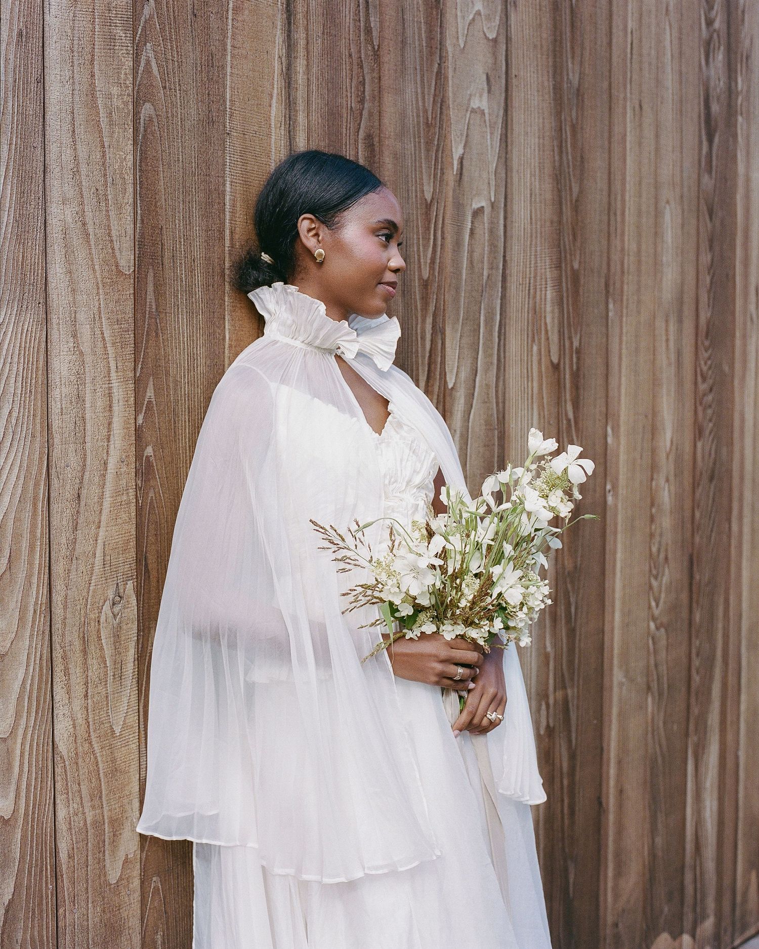 A Black bride in a white chiffon gown and cape holds a delicate white floral bouquet against a wooden wall.