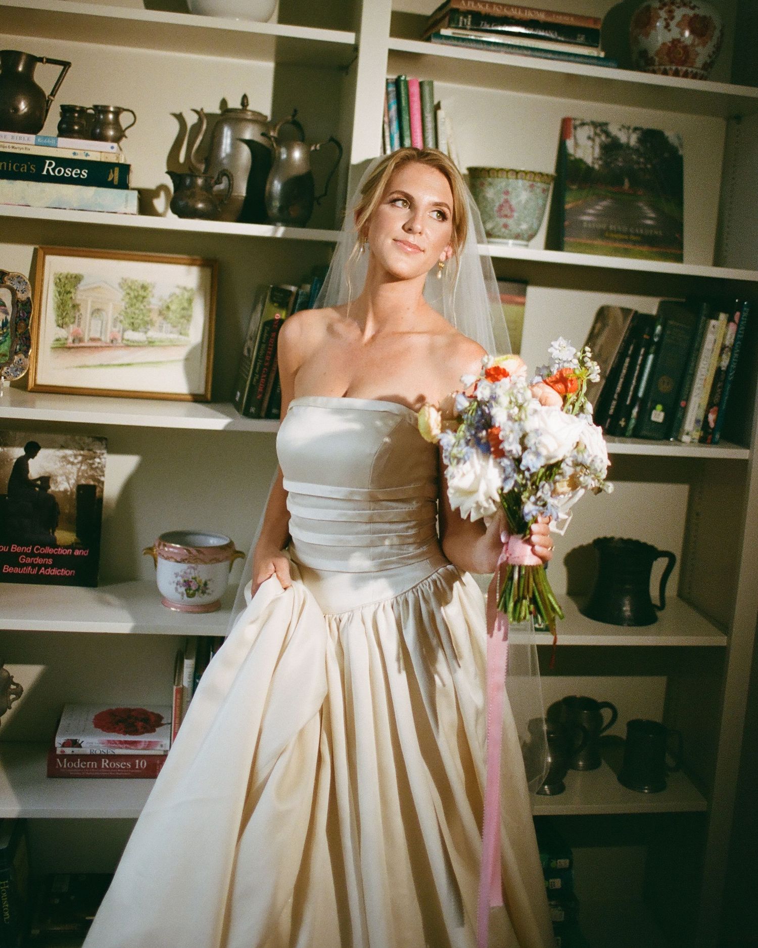 An elegant cream-colored wedding dress with a strapless neckline and full skirt is shown in a library setting.