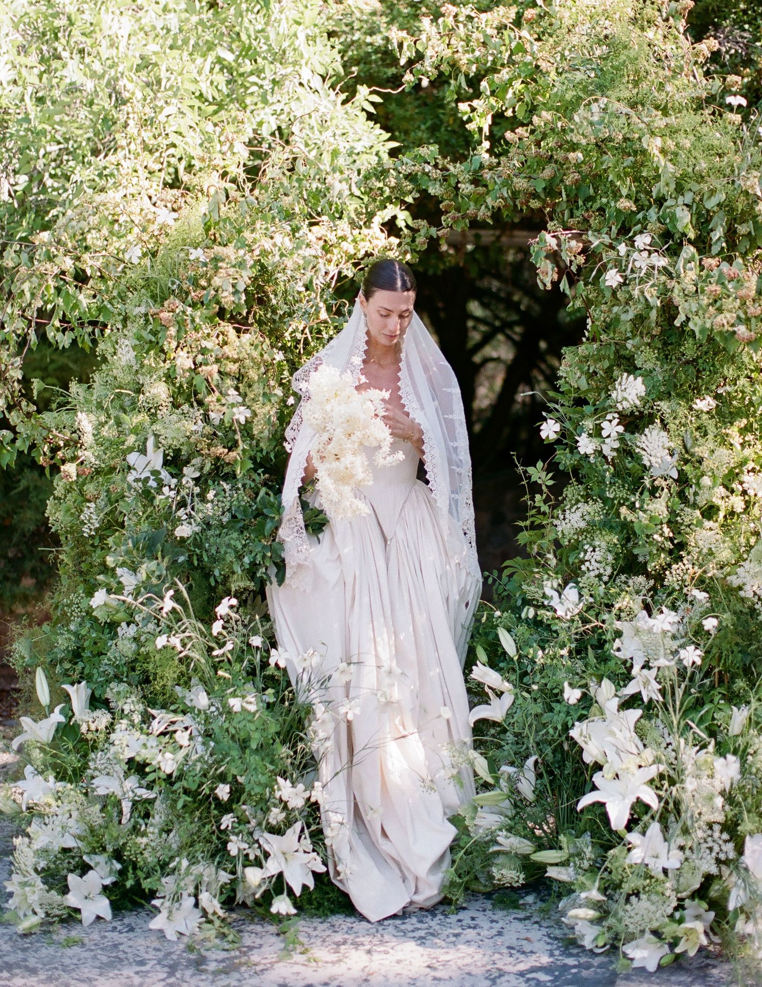 Bride in flowing white gown and veil holding bouquet, surrounded by lush white florals and greenery outdoors.