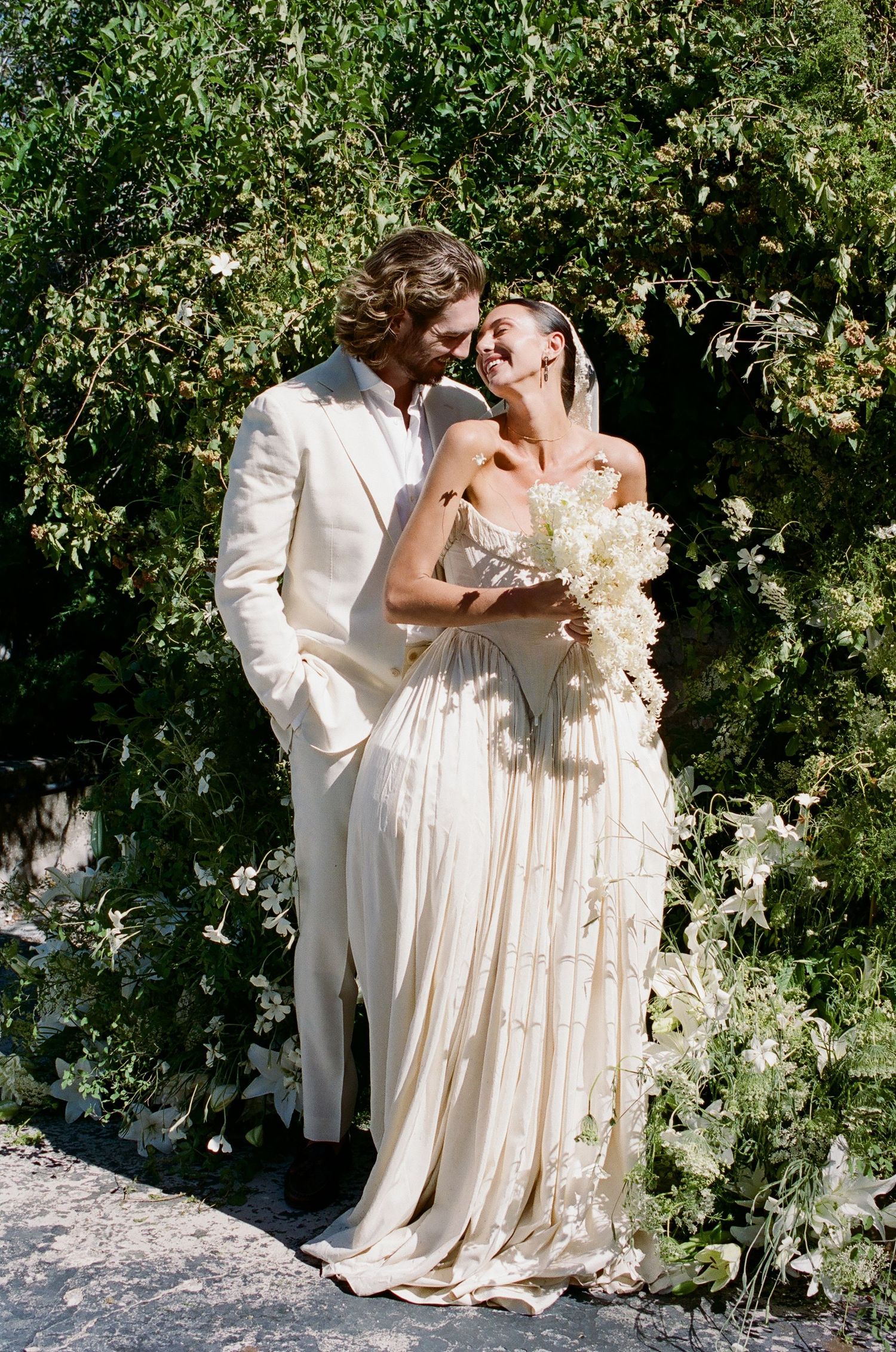 A bride and groom laugh together in front of lush greenery, she holds white flowers in a flowing cream gown.
