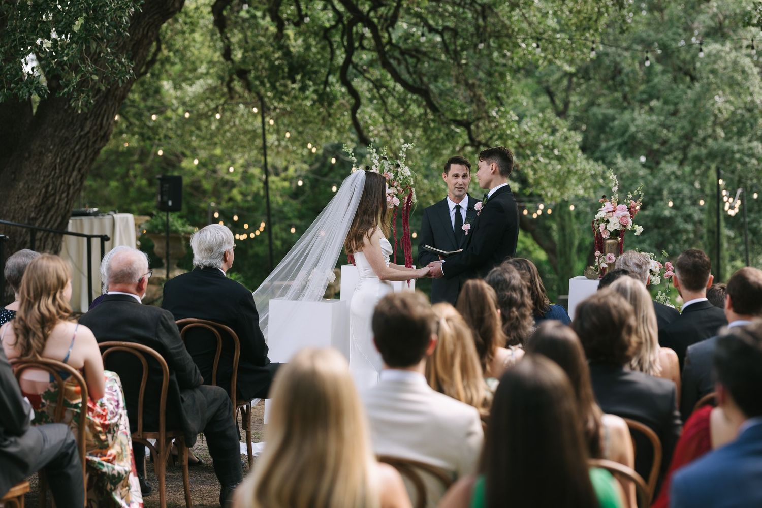 Wide view of outdoor wedding ceremony with guests seated under large trees.