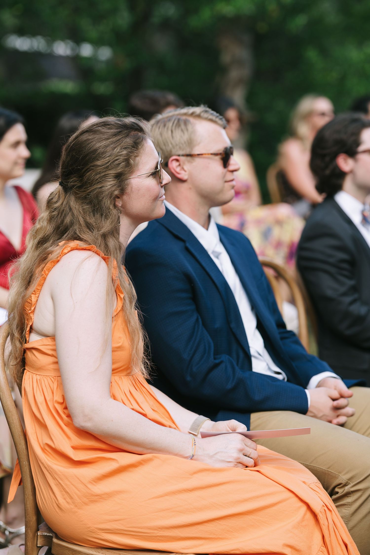 Wedding guests in formal attire seated on wooden chairs at outdoor garden ceremony.
