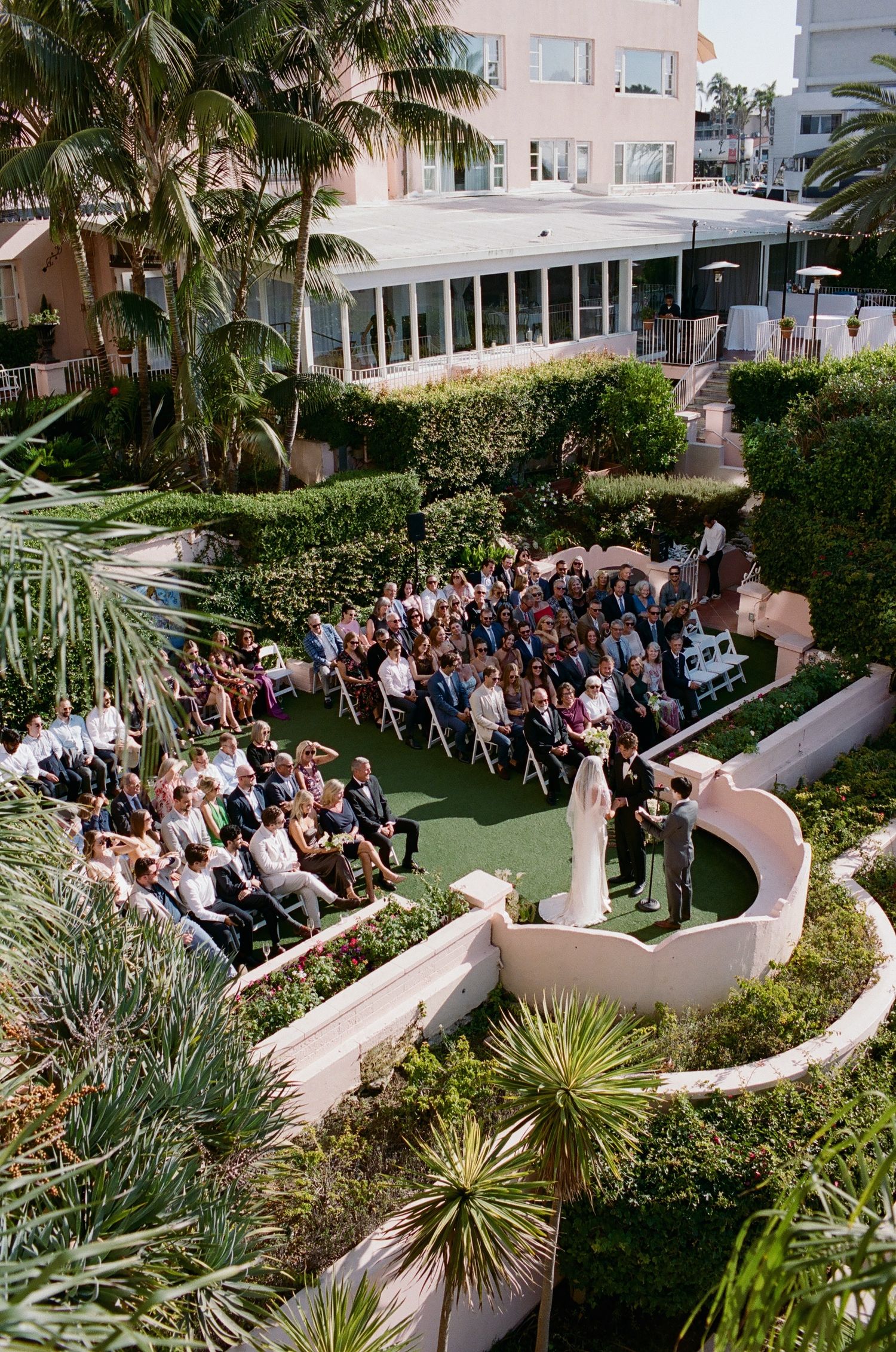 Aerial view of a wedding ceremony in a tropical garden with curved white walls and palm trees.