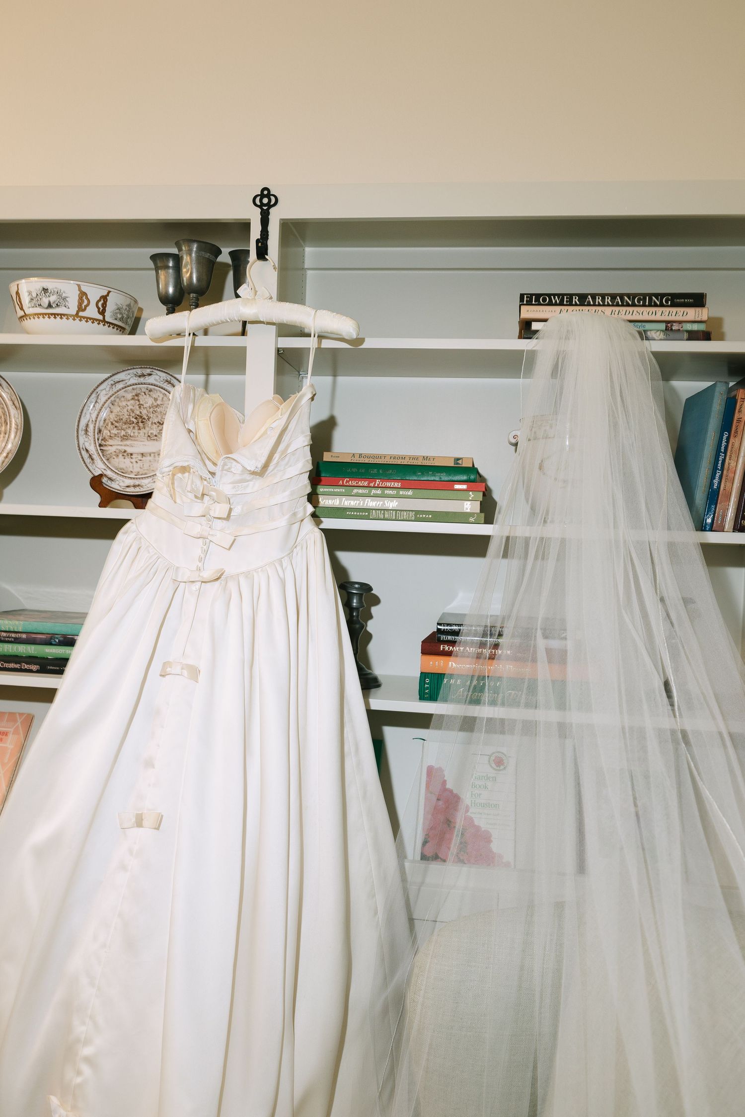White wedding dress with tulle veil displayed on hangers against a bookshelf.