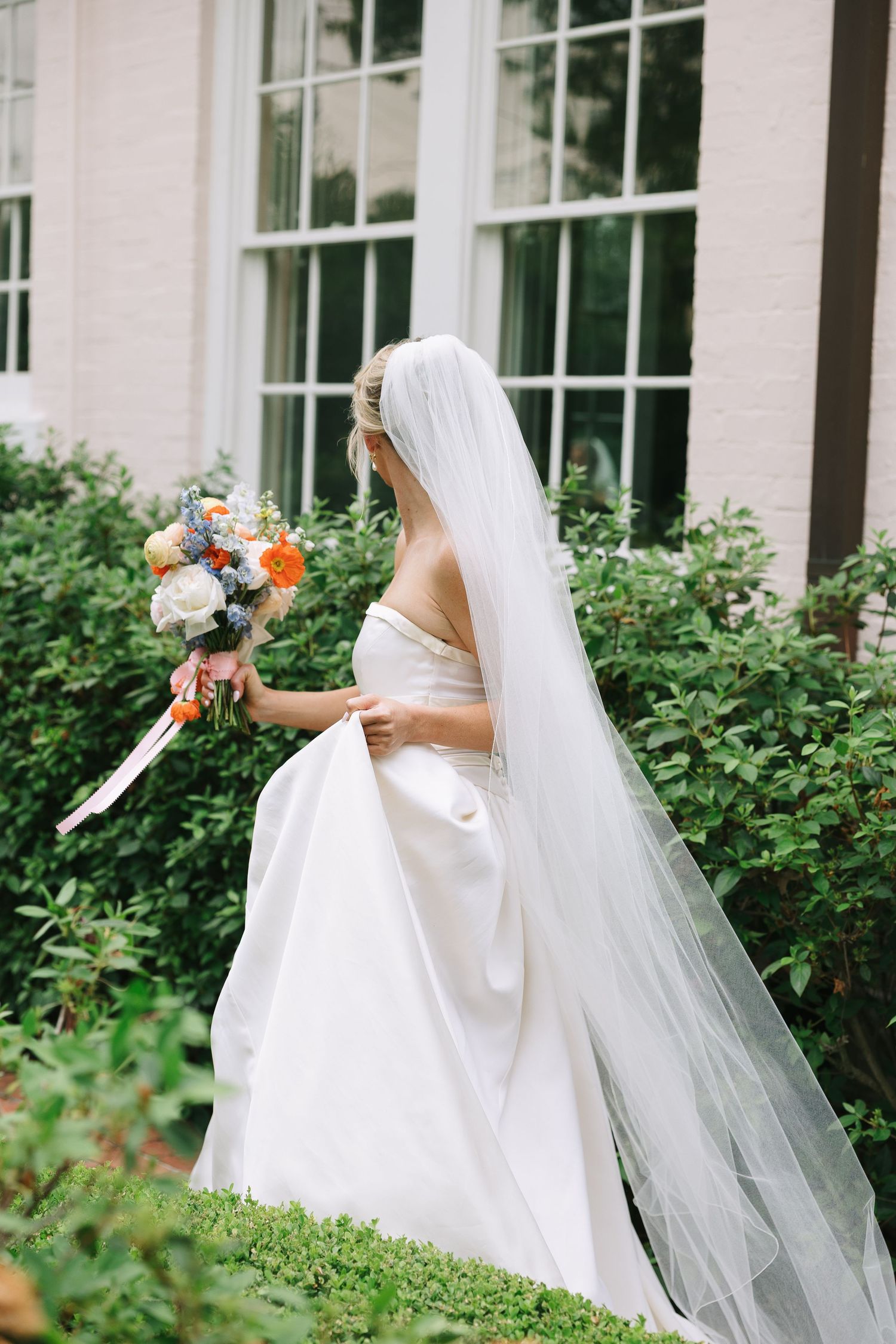 Elegant bride in flowing white dress and veil walking through garden setting.