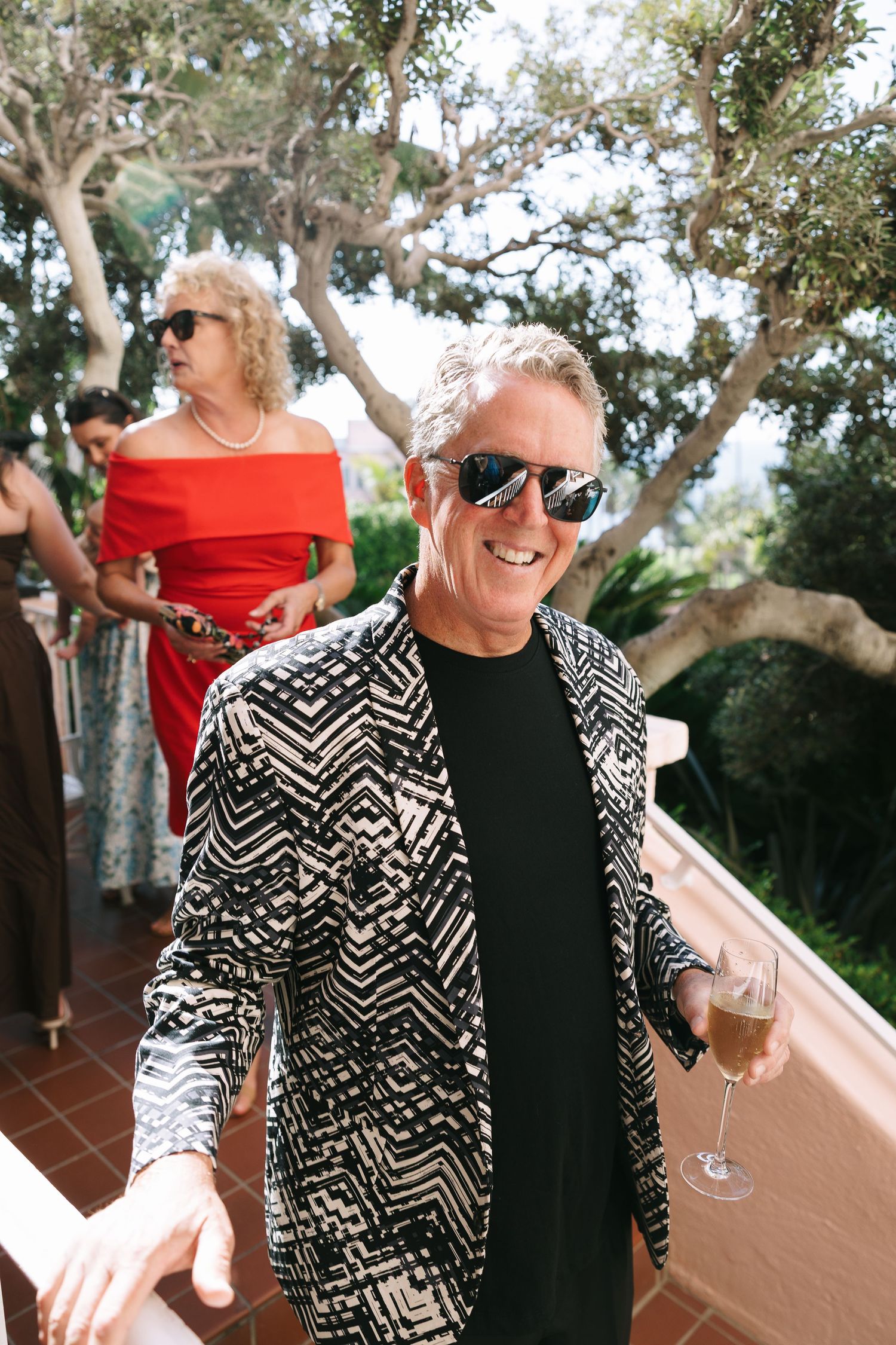 Wedding guest wearing black and white patterned jacket and sunglasses dances at outdoor celebration.