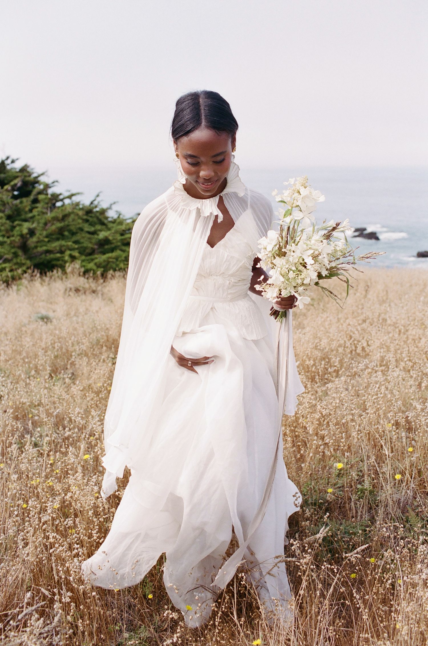 Bride in white gown and cape holding bouquet in golden field with ocean backdrop.