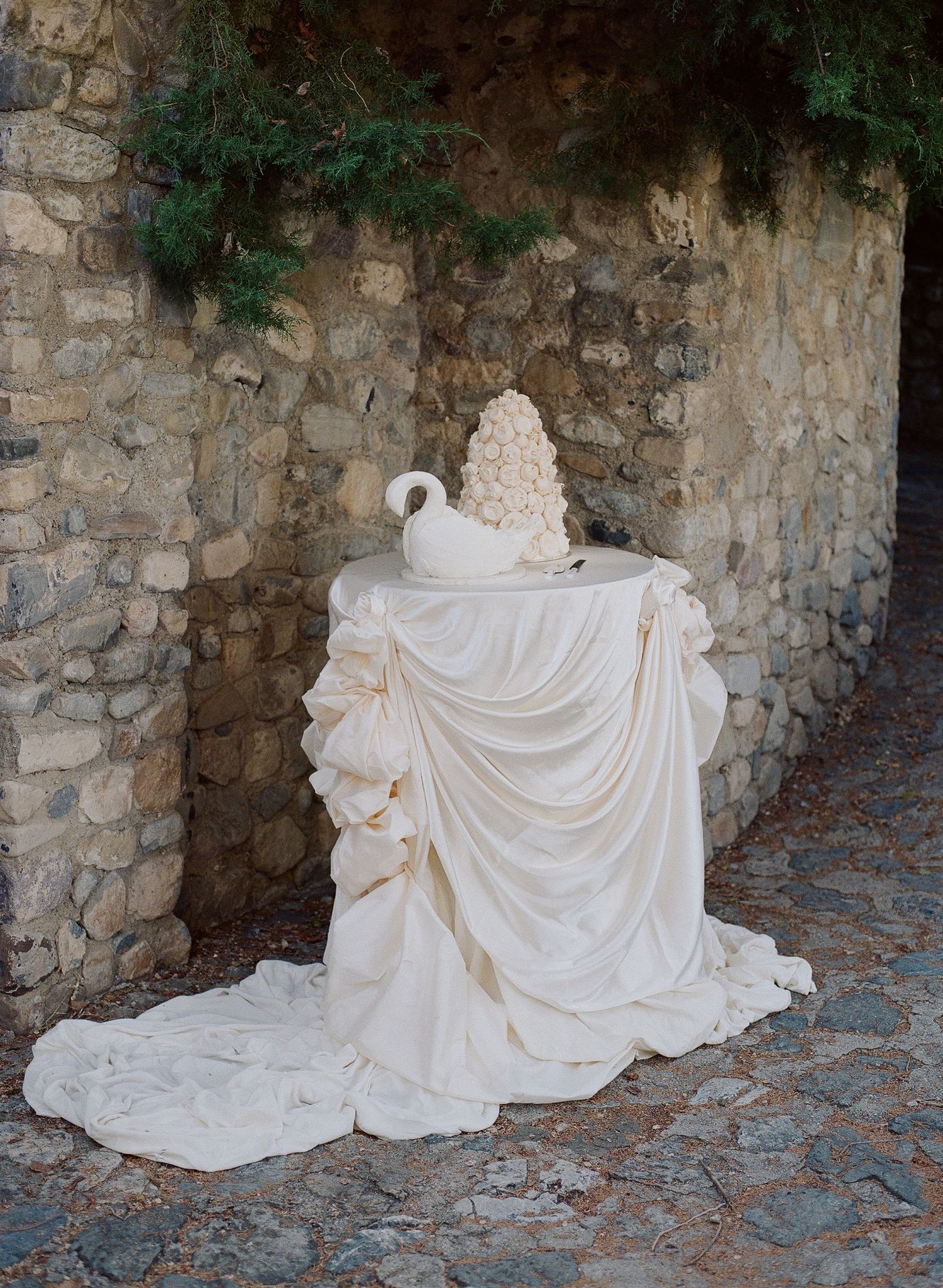 Elegant wedding cake display on draped white fabric table against rustic stone wall with greenery backdrop.