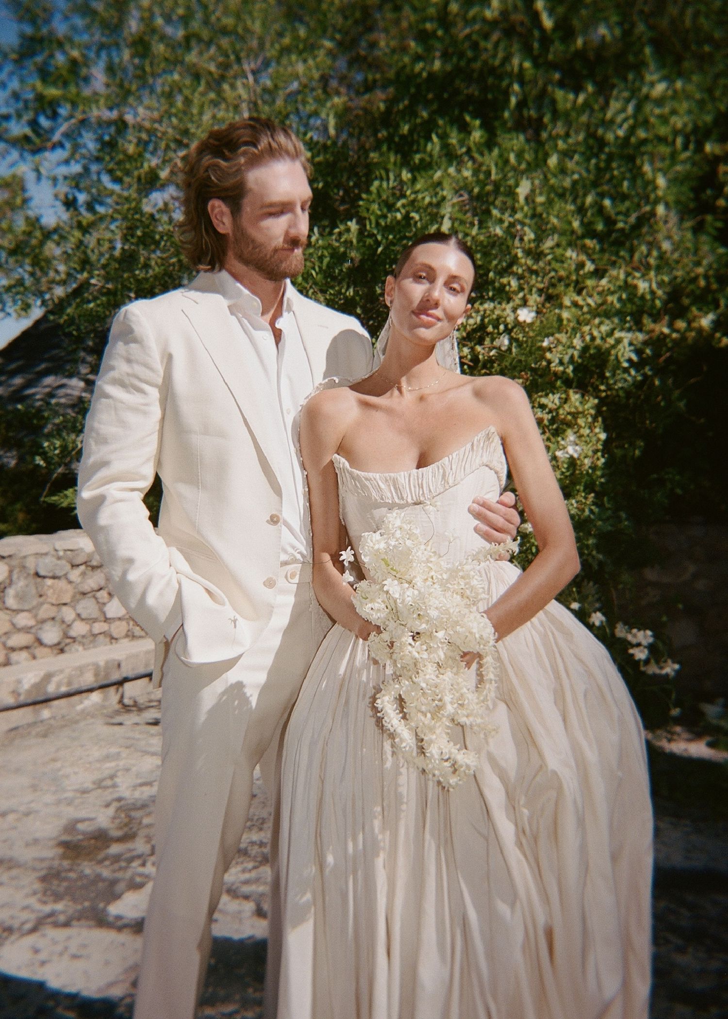 A couple poses outdoors in elegant wedding attire, the bride holding a white bouquet in a strapless gown beside a groom in a cream suit.