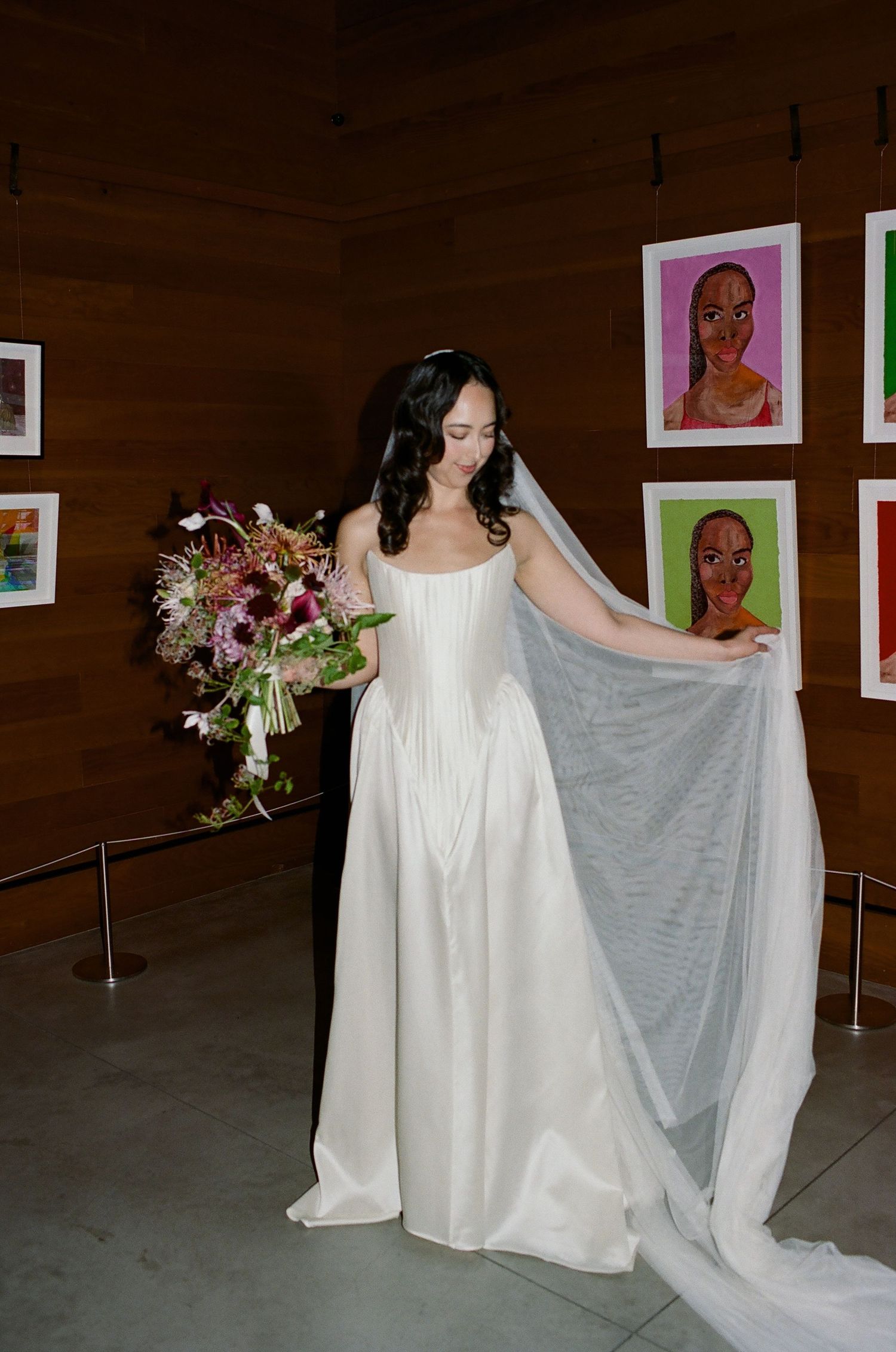 A figure in a flowing white dress and veil stands in front of a gallery wall displaying colorful portrait artwork.