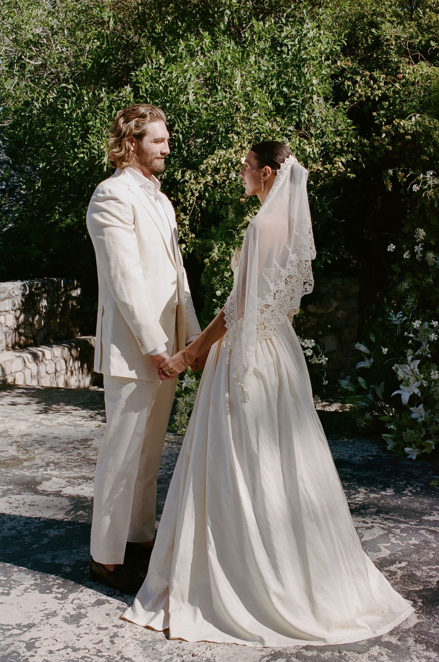 A bride and groom in ivory wedding attire hold hands and face each other outdoors amid lush green trees.