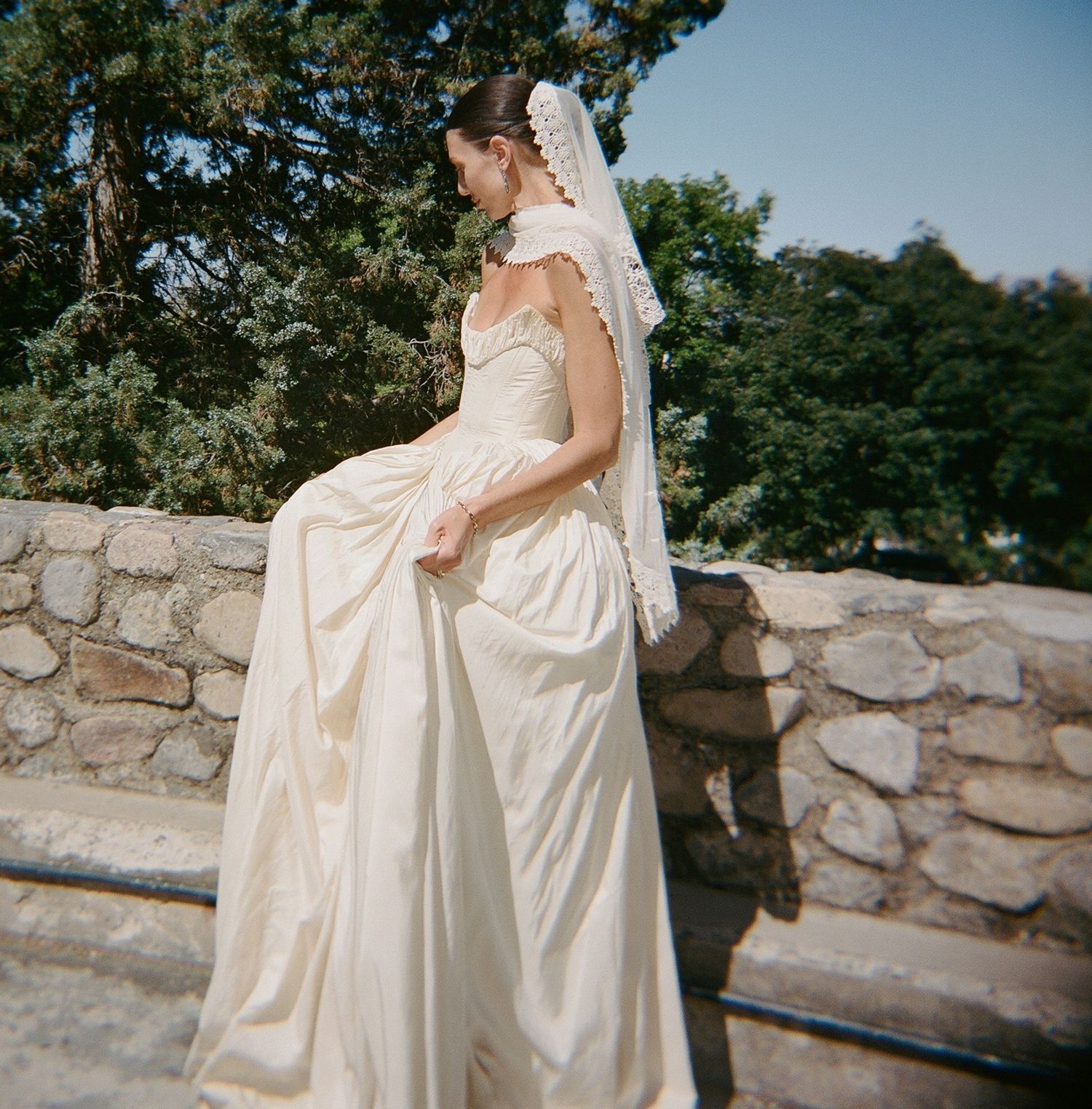 Bride in ivory gown and lace veil sits on stone wall surrounded by lush green trees on a sunny day.