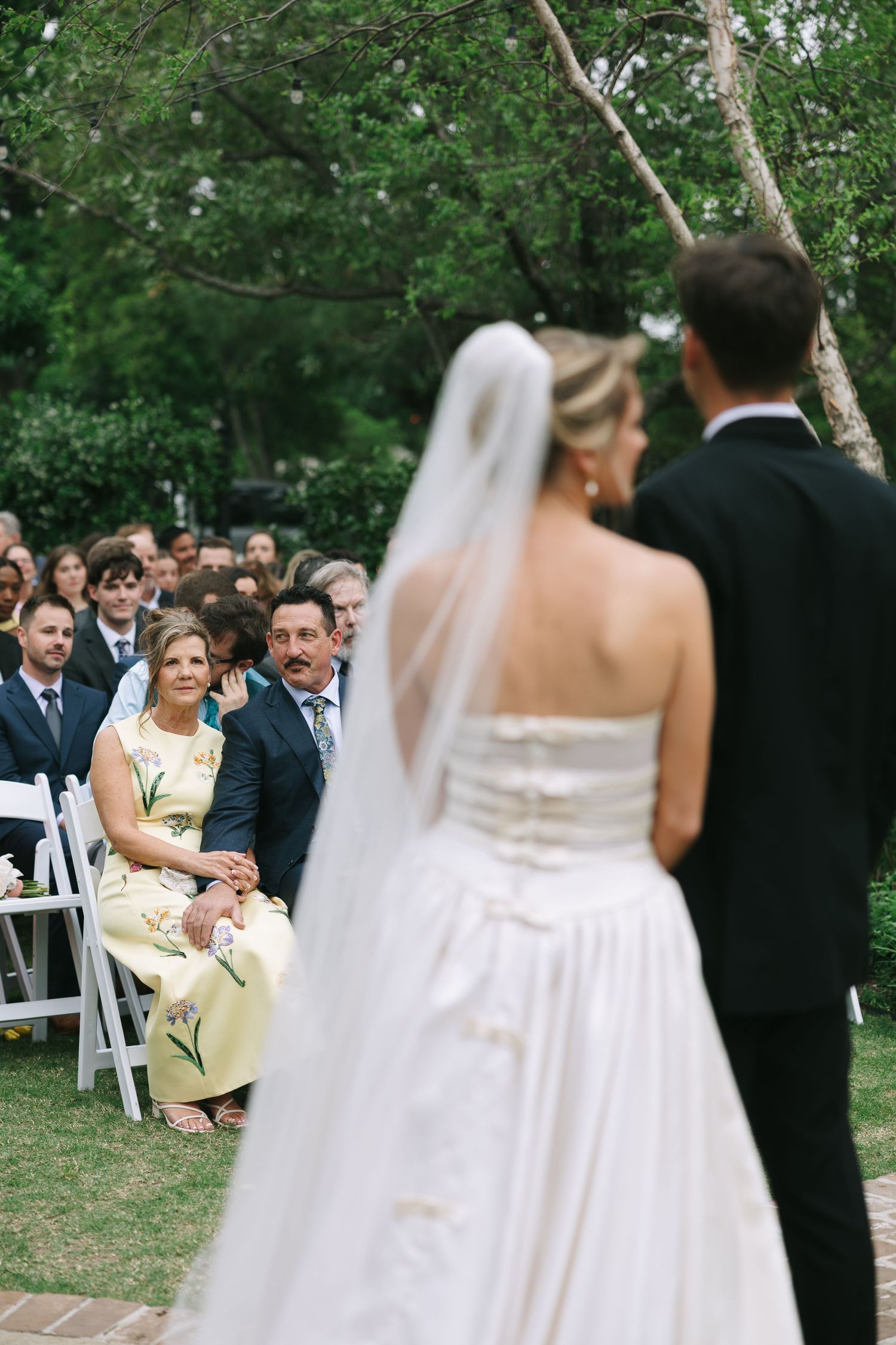 Wedding ceremony in garden setting with guests seated on white chairs.