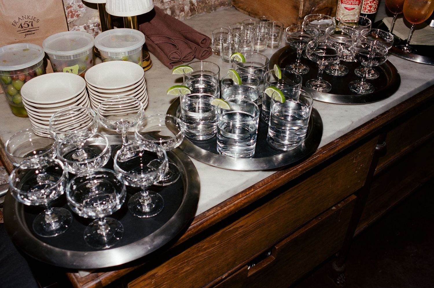 Stacks of white plates and wine glasses arranged on black serving trays on a wooden sideboard.