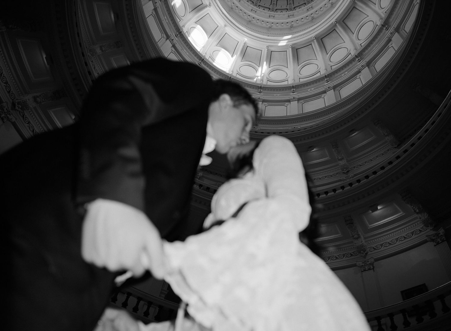 Black and white photo of a bride and groom sharing a kiss inside a grand domed building with ornate architecture.