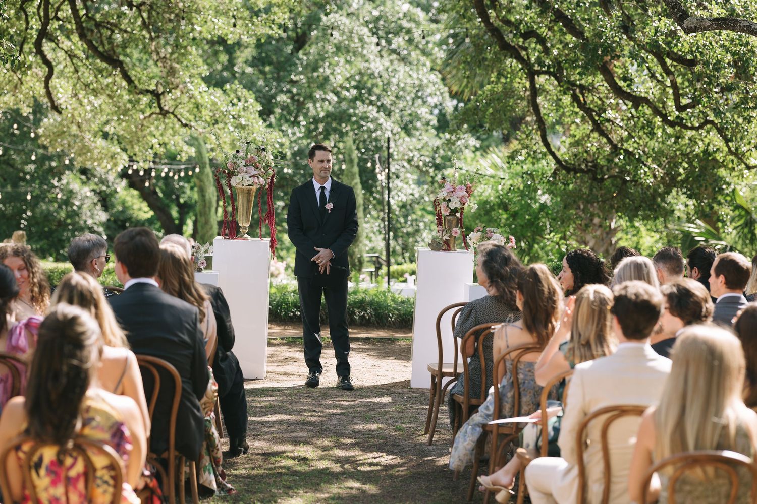 Outdoor wedding ceremony setup with guests seated under large trees.