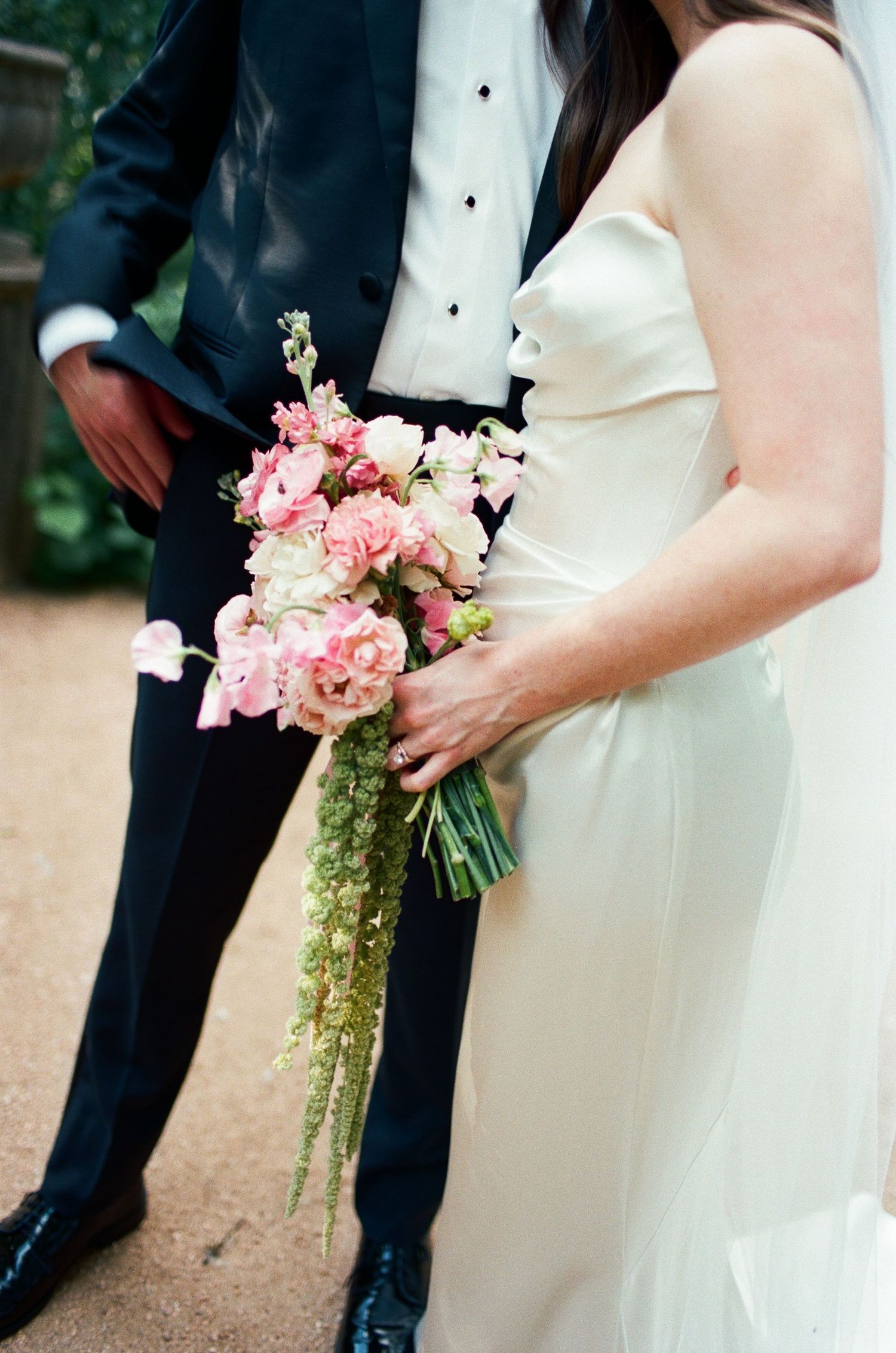 A pink and white wedding bouquet held between formal attire at an outdoor ceremony.