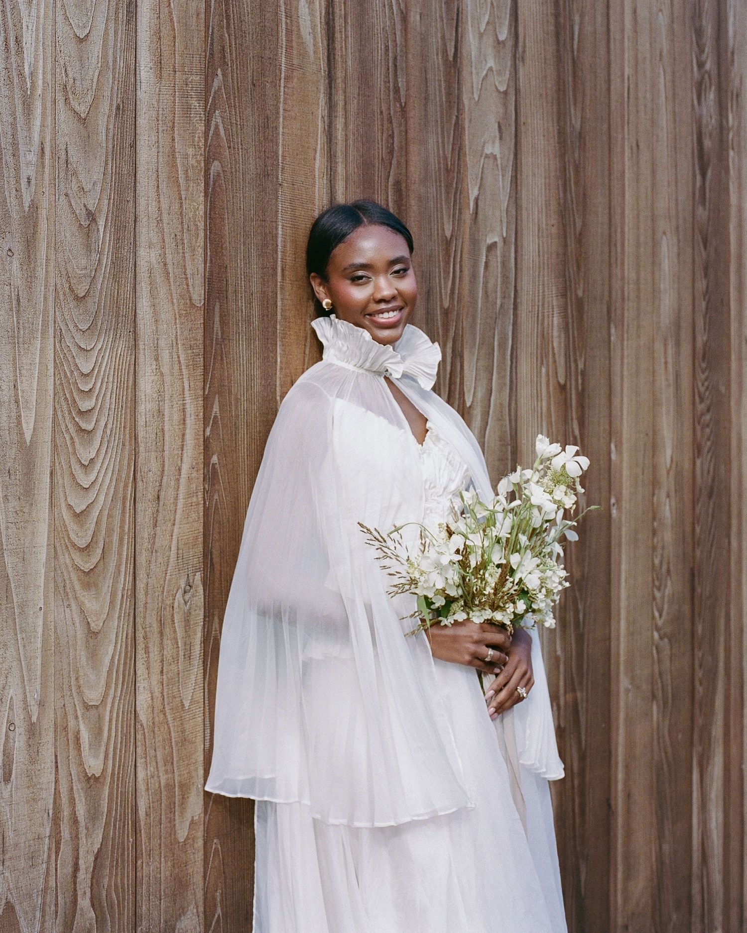 Smiling bride in white chiffon cape gown holding wildflower bouquet against rustic wood backdrop.