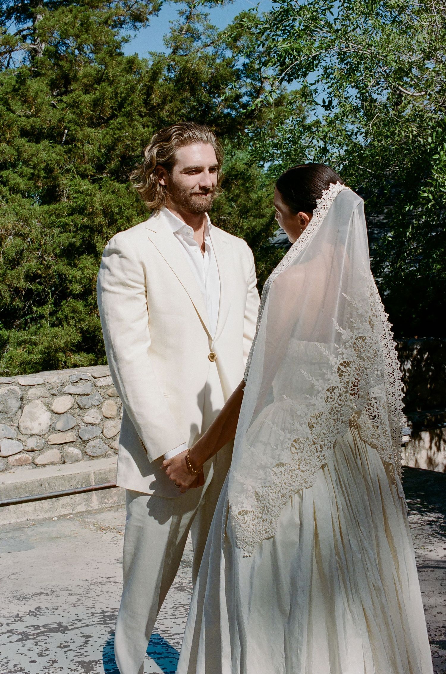 A couple in matching ivory outfits holds hands outdoors, the bride wearing an embroidered veil and the groom in a white suit.