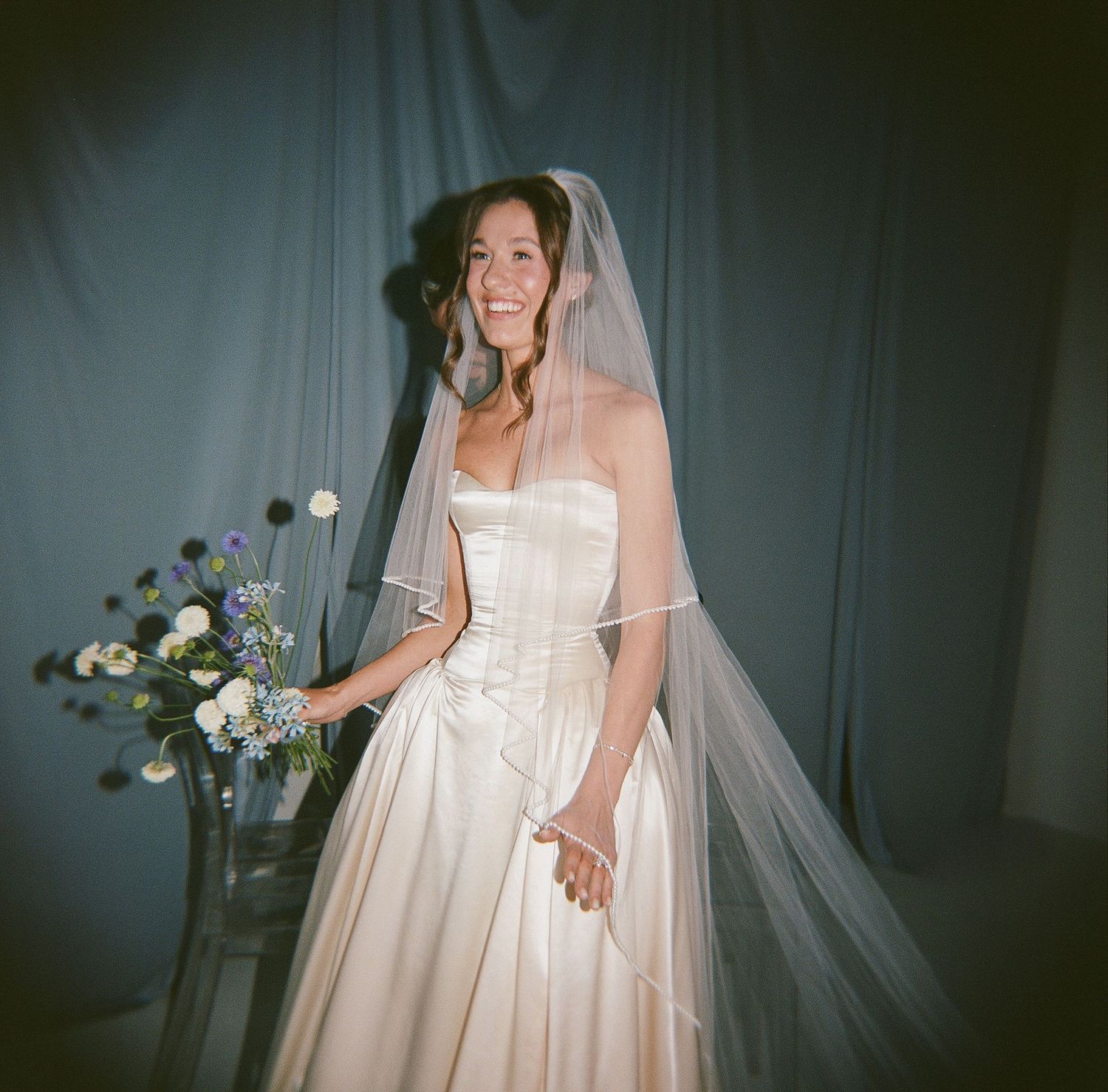 Smiling bride in strapless satin wedding gown with tulle veil, posing beside blue floral arrangement on gray backdrop.