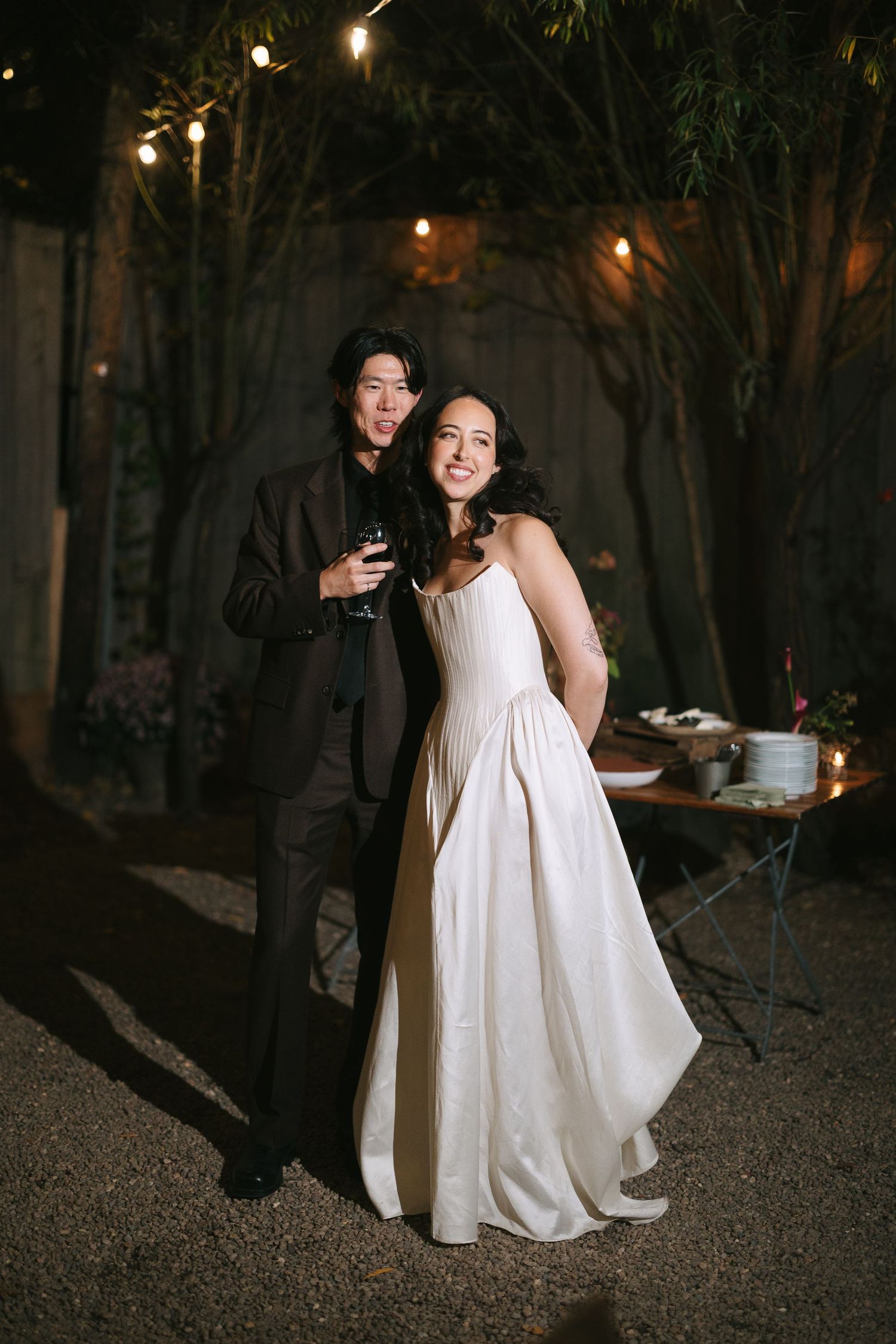 A couple in formal wedding attire poses together outdoors at night under string lights.
