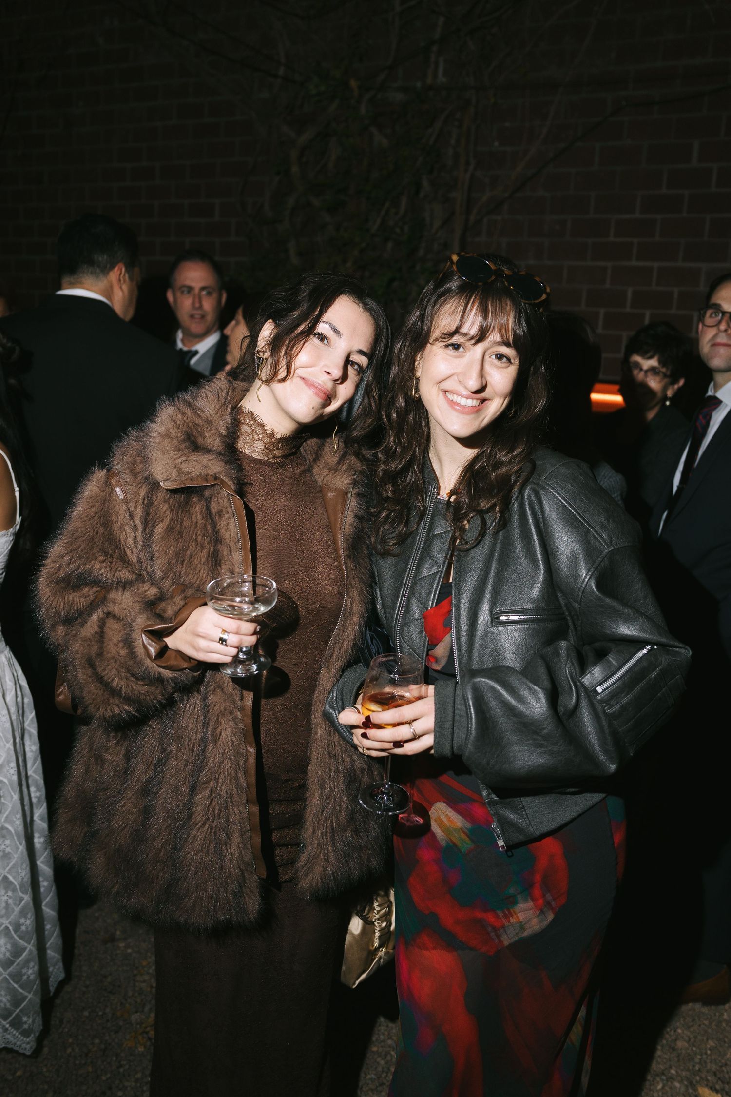 Party guests in winter coats mingling and holding drinks at a dimly lit gathering.