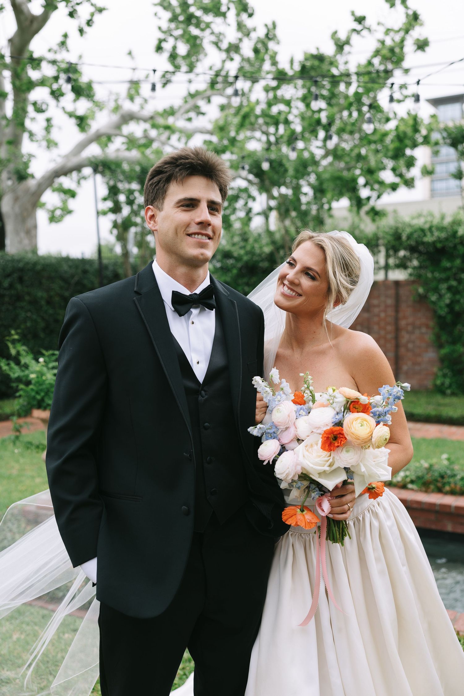 Wedding couple shares a candid moment in a garden with lush greenery and brick walls.
