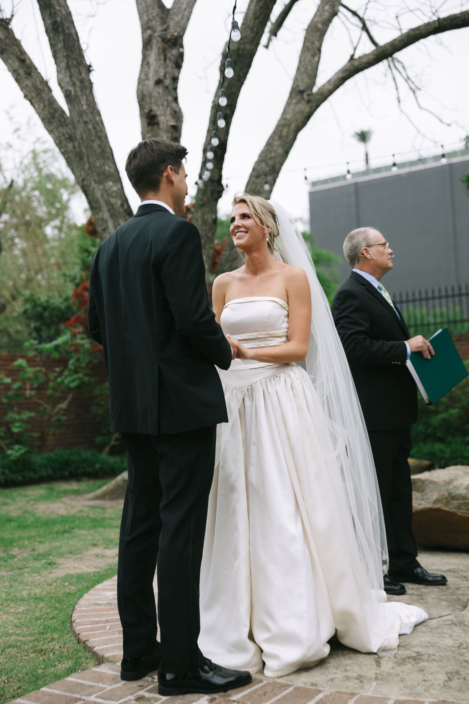Elegant wedding procession walking along a garden path with formal attire and flowing white dress.