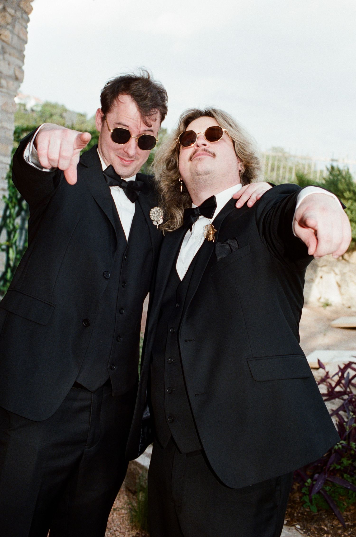 Two men in black tuxedos and sunglasses pointing at camera outdoors.