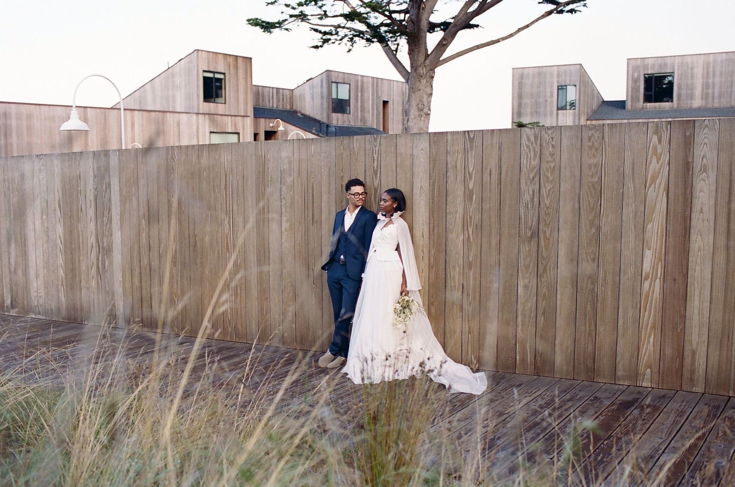 A bride and groom stand together by a wooden fence surrounded by tall grass and modern cedar buildings on their wedding day.