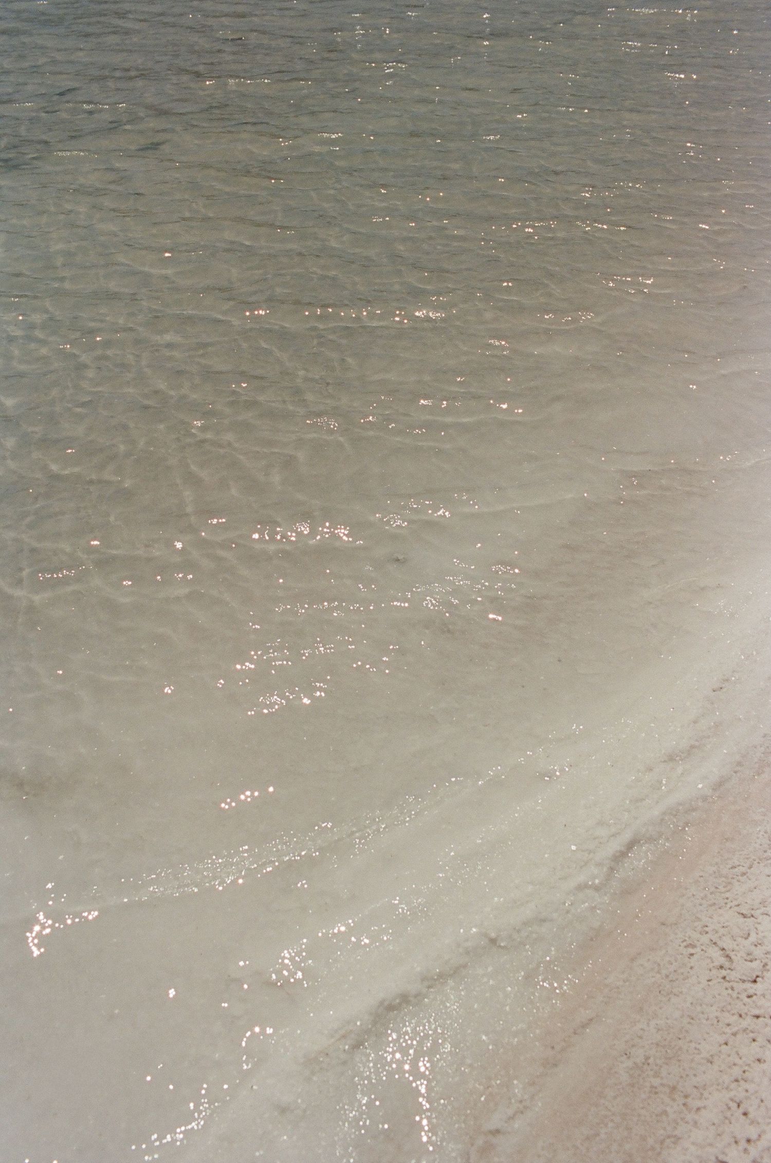 Shallow clear ocean waves gently washing over a sandy beach shore with sunlight sparkling on the water surface.