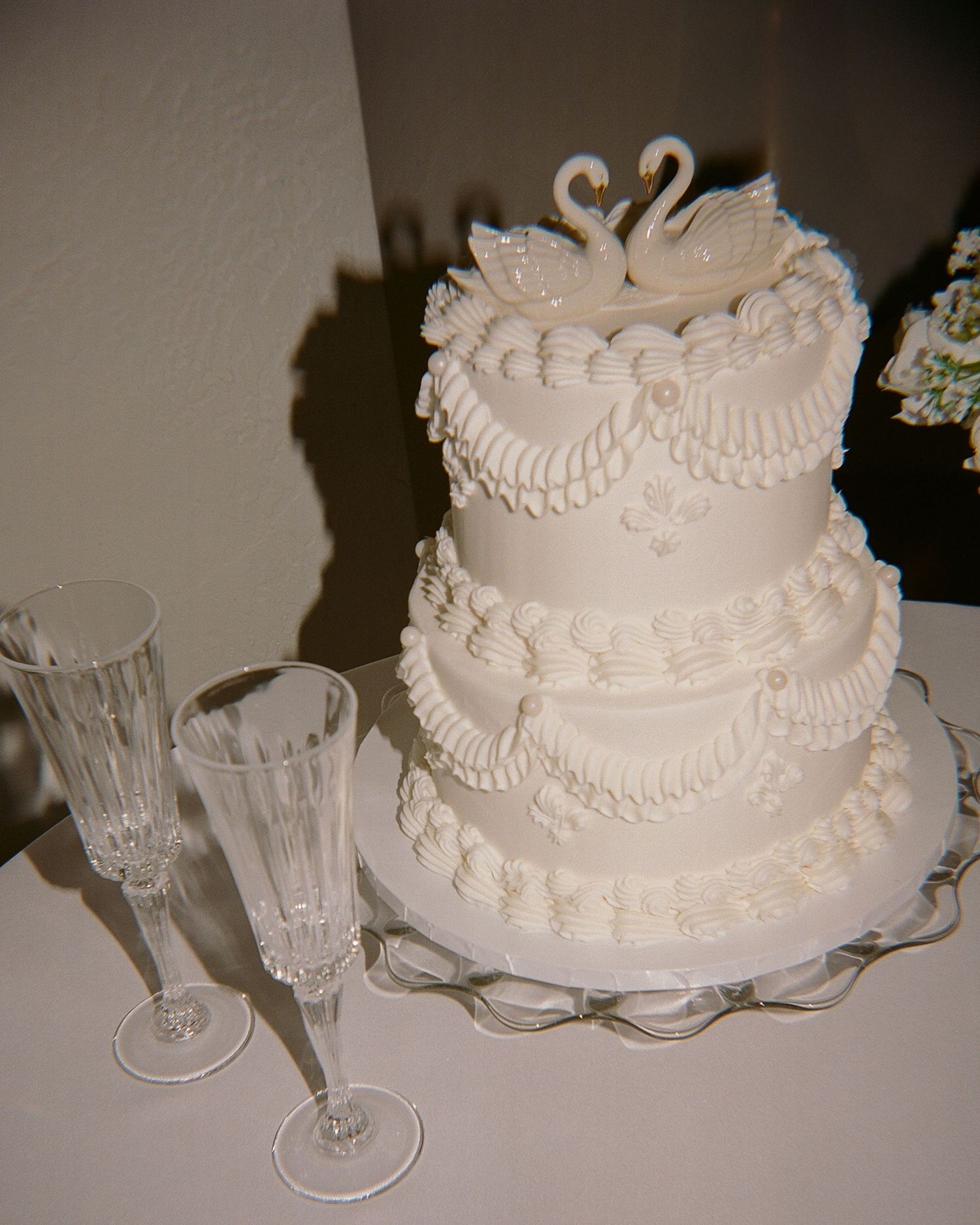 A white tiered wedding cake with delicate piped frosting details sits on a glass cake stand next to crystal champagne flutes.