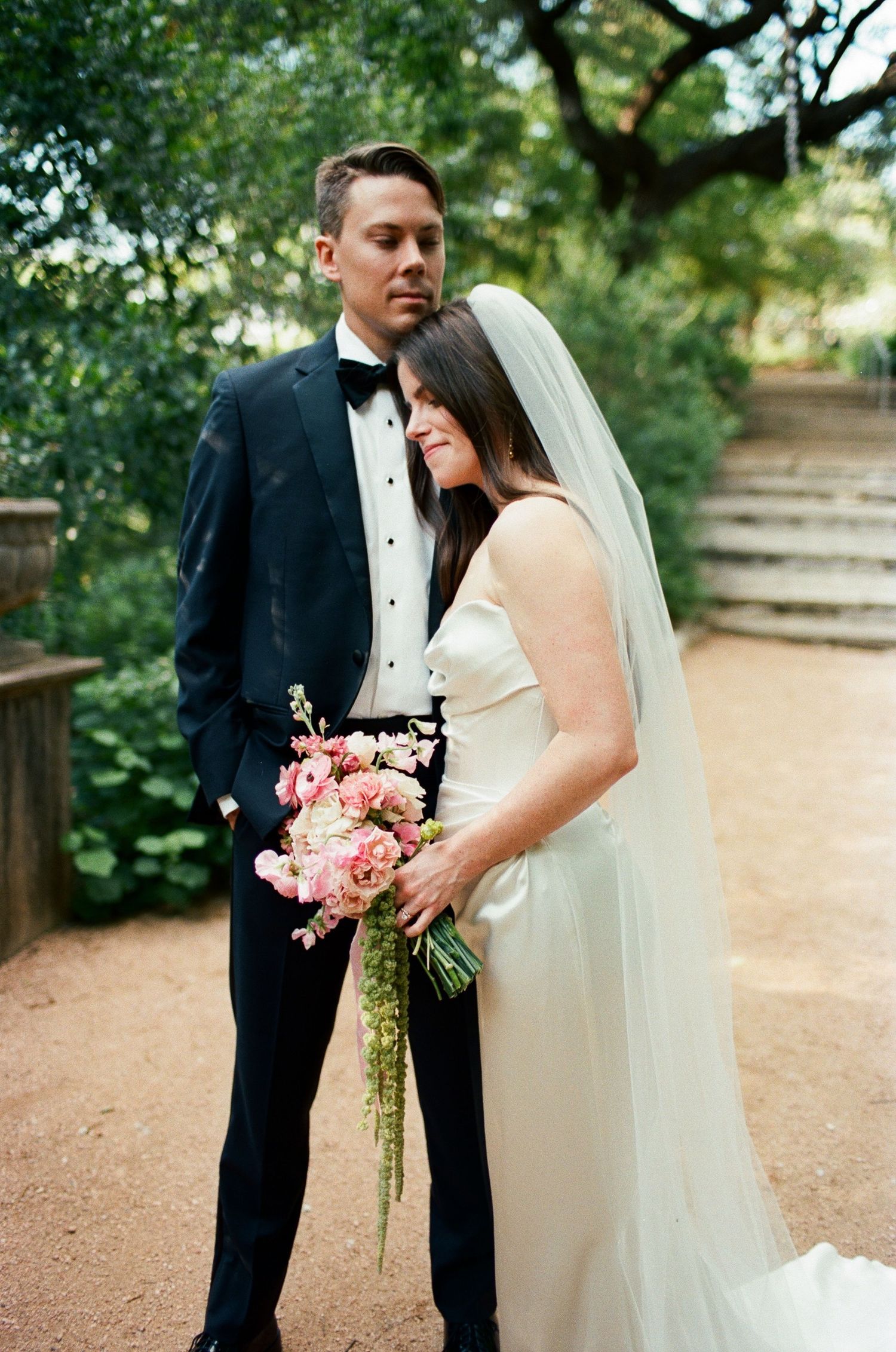 Elegant wedding portrait on garden path with pink bouquet and flowing veil.