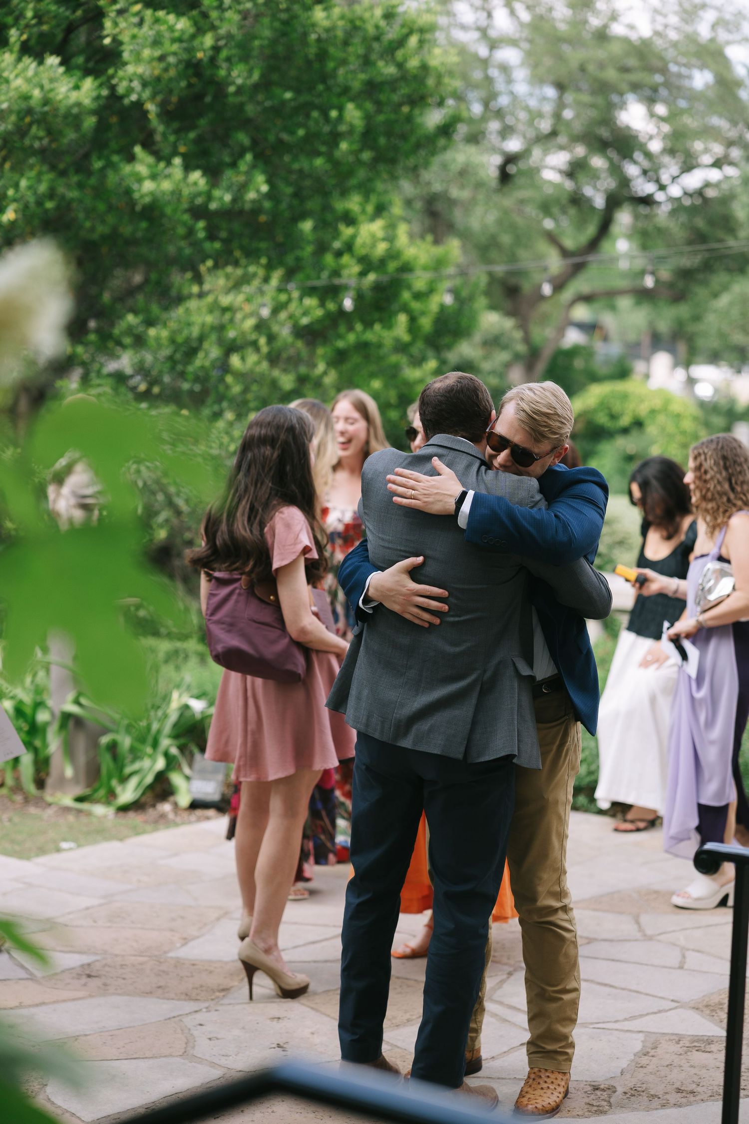 Wedding guests embrace and hug each other on a stone path surrounded by greenery.