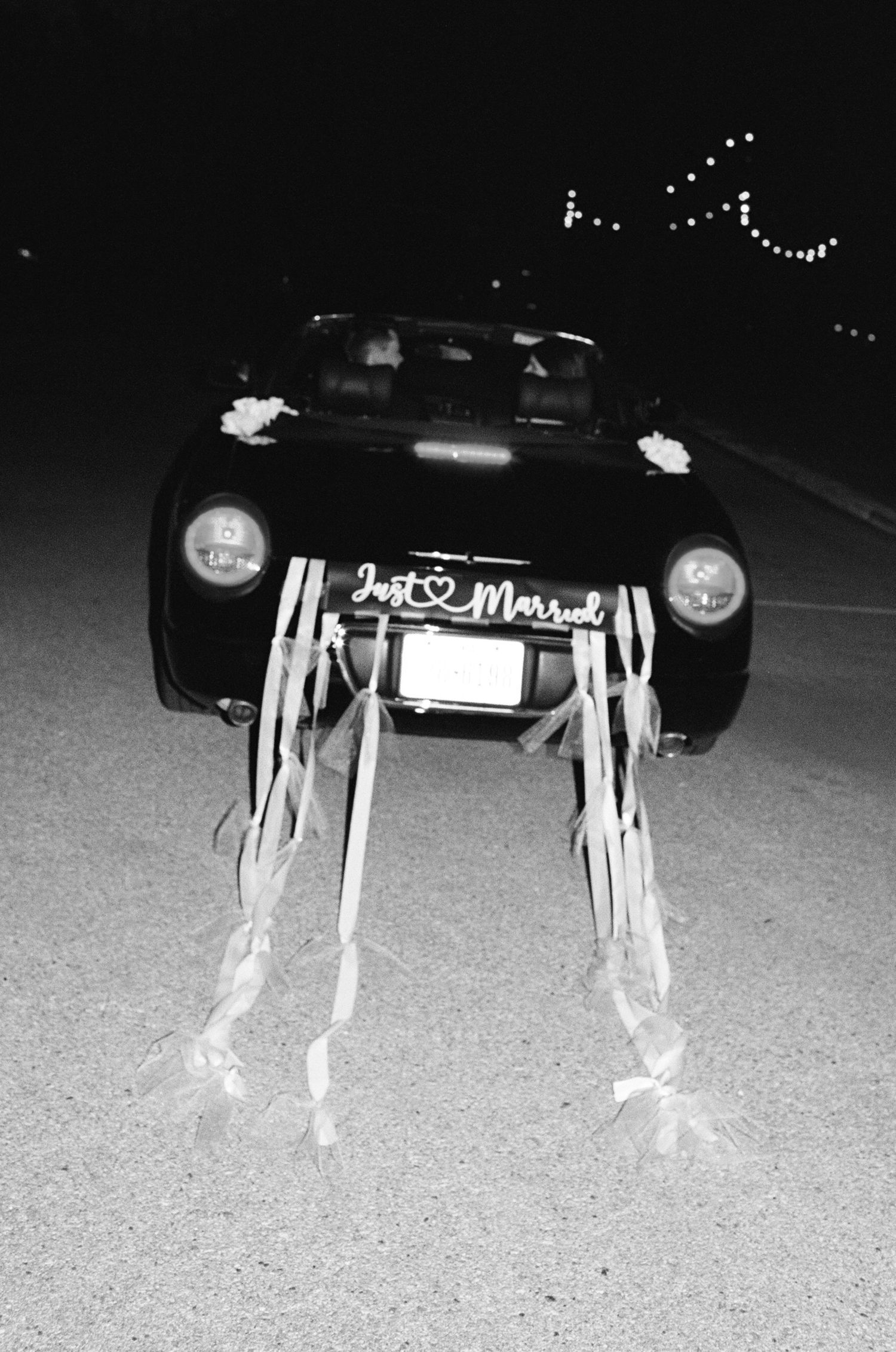 Black and white image of a decorated wedding getaway car with streamers at night.